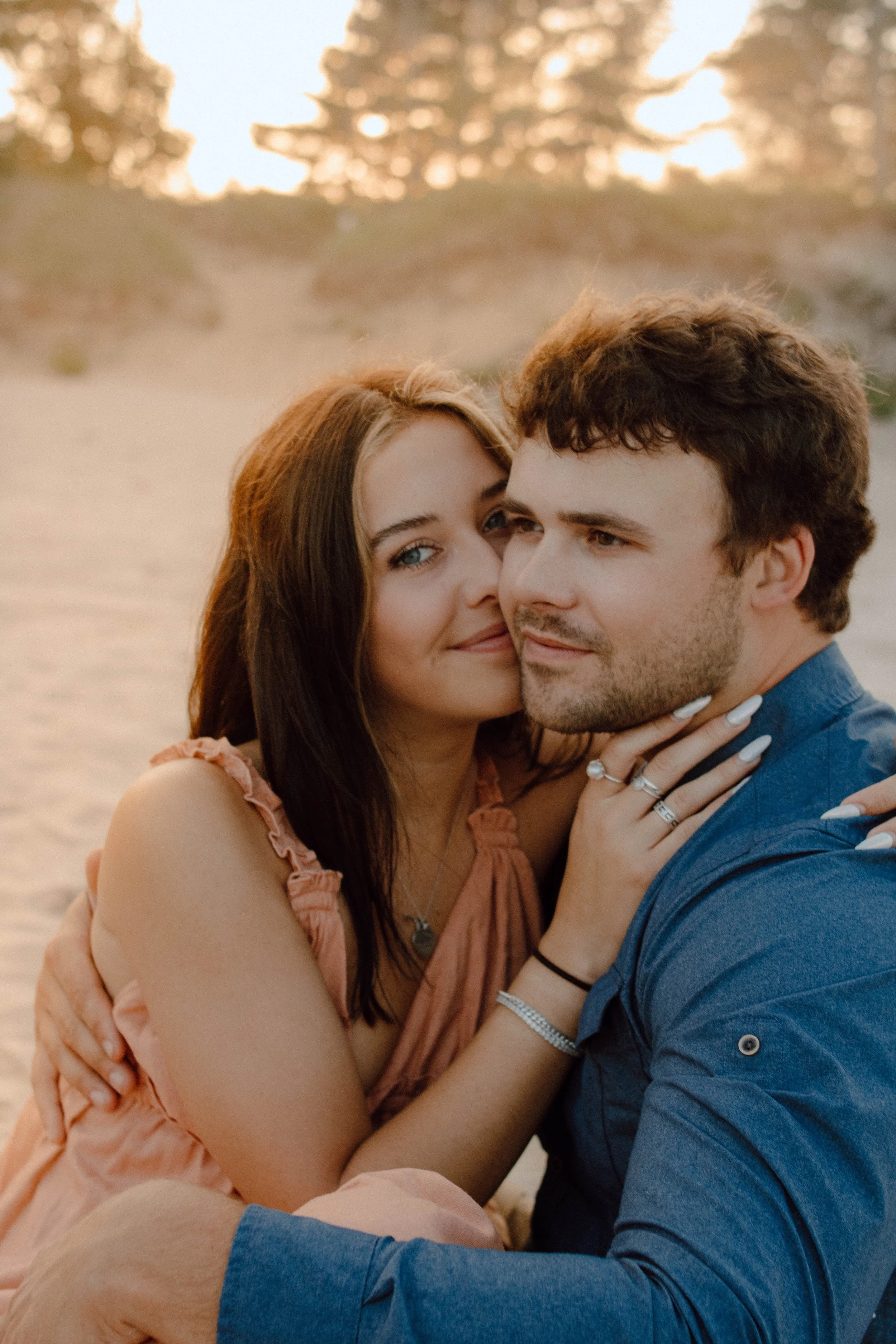 A couple hugging on a sandy beach during sunset, with trees in the background.
