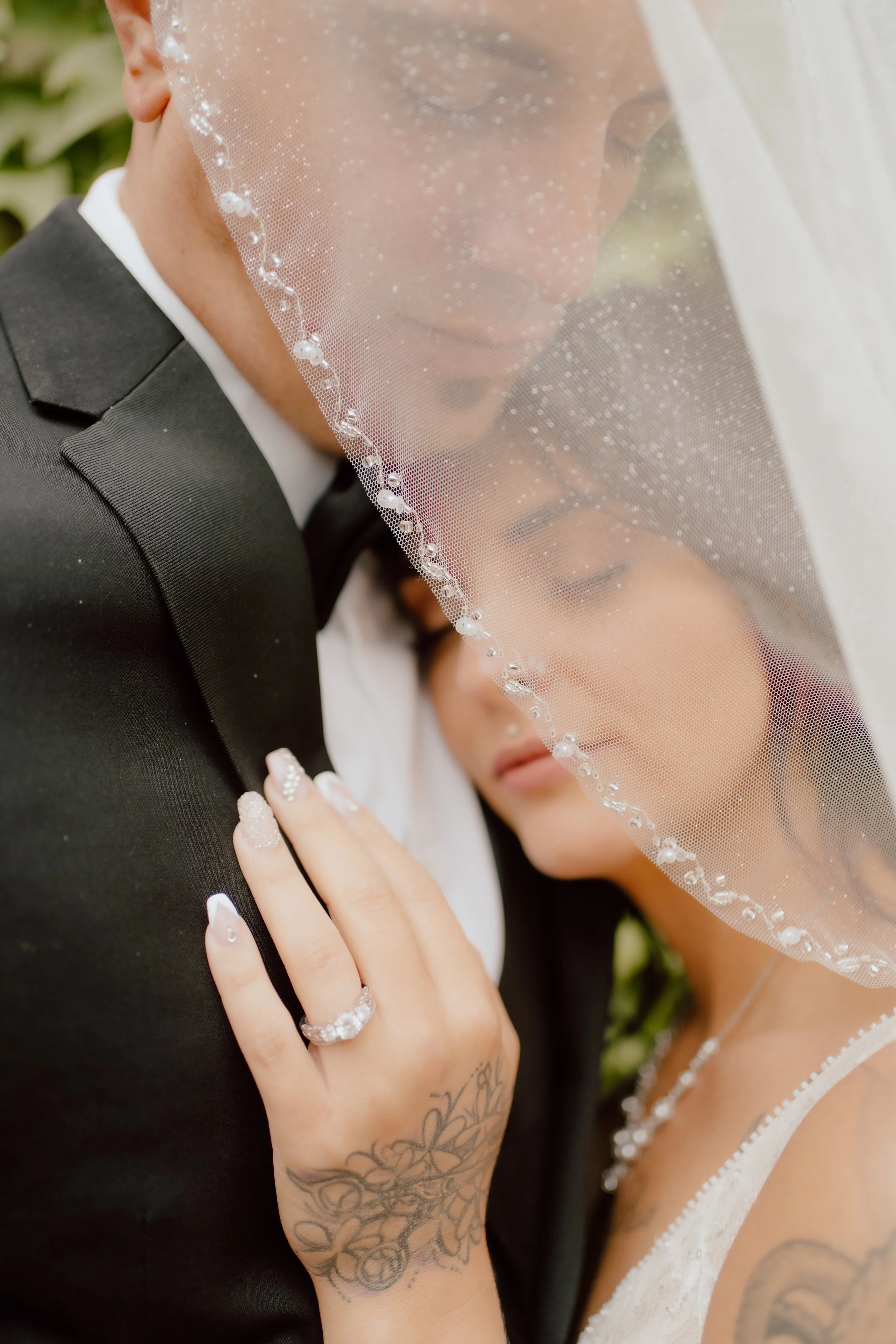 A close-up of a bride and groom sharing an intimate moment, with the bride resting her head on the groom’s shoulder. The bride wears a veil with delicate beadwork and has tattoos on her hand and arm. The groom is dressed in a black tuxedo. The scene 