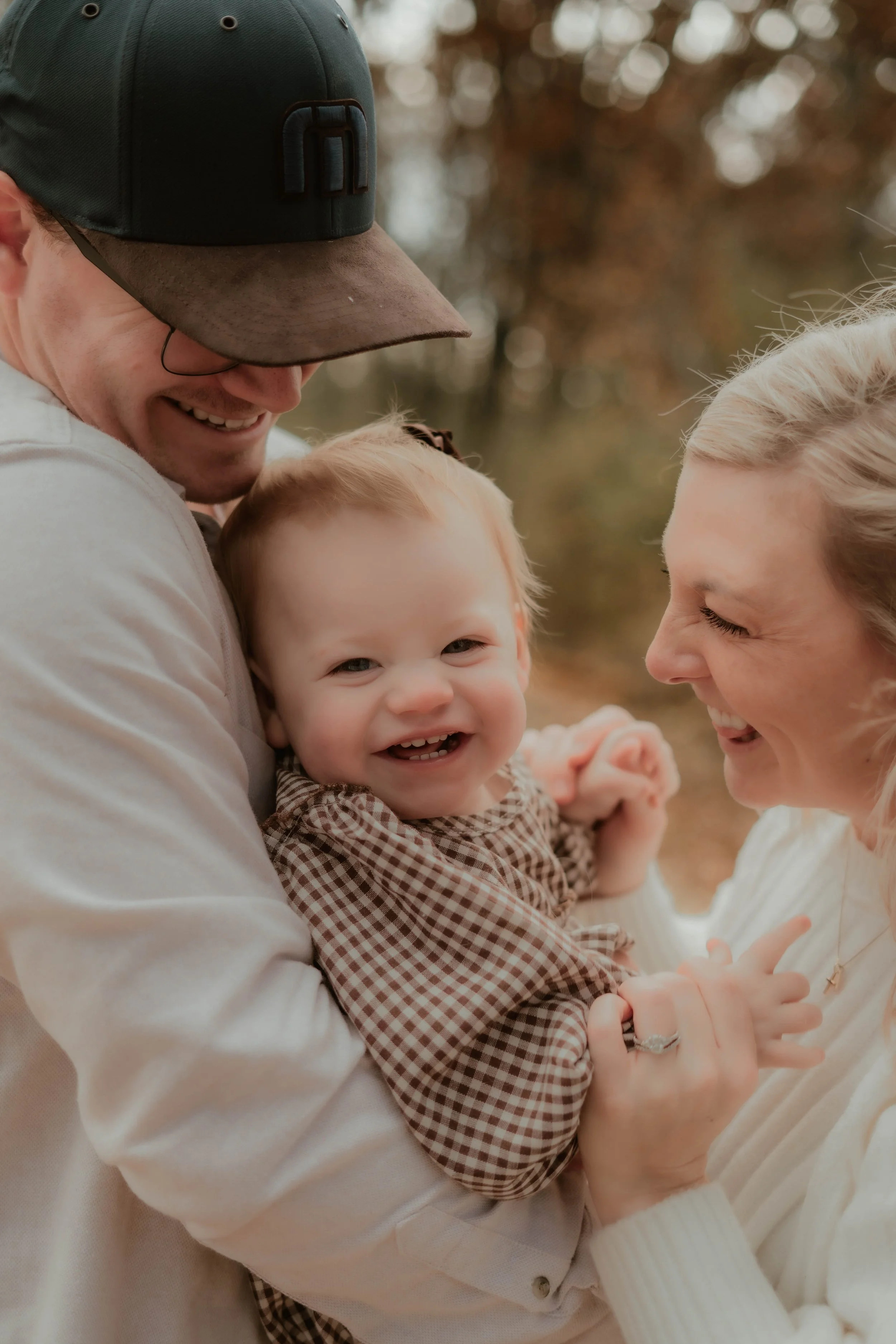 Smiling family outdoors, a man and woman holding a happy toddler, fall foliage in background.