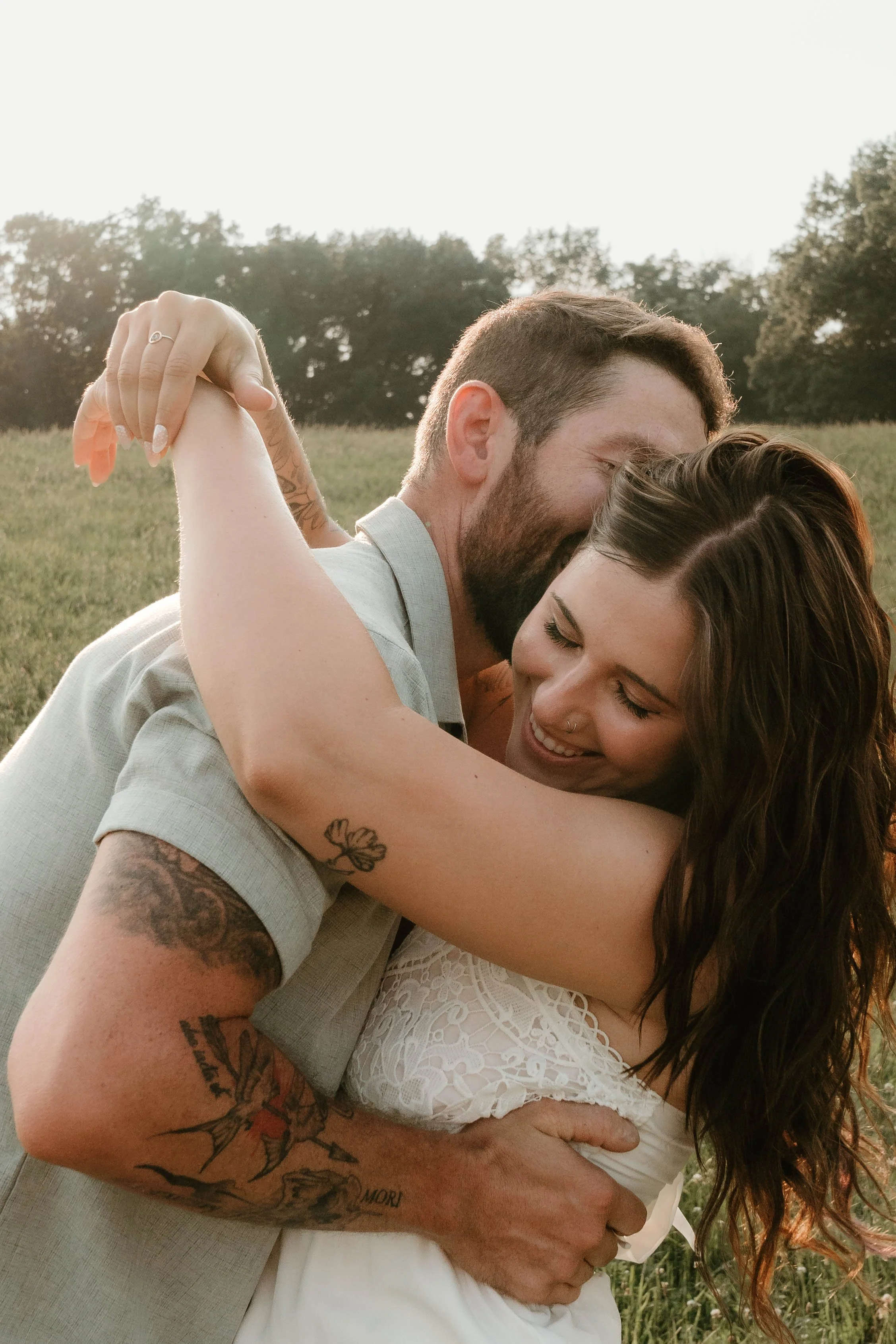 A couple embracing and smiling outdoors during sunset, with trees and open field in the background.