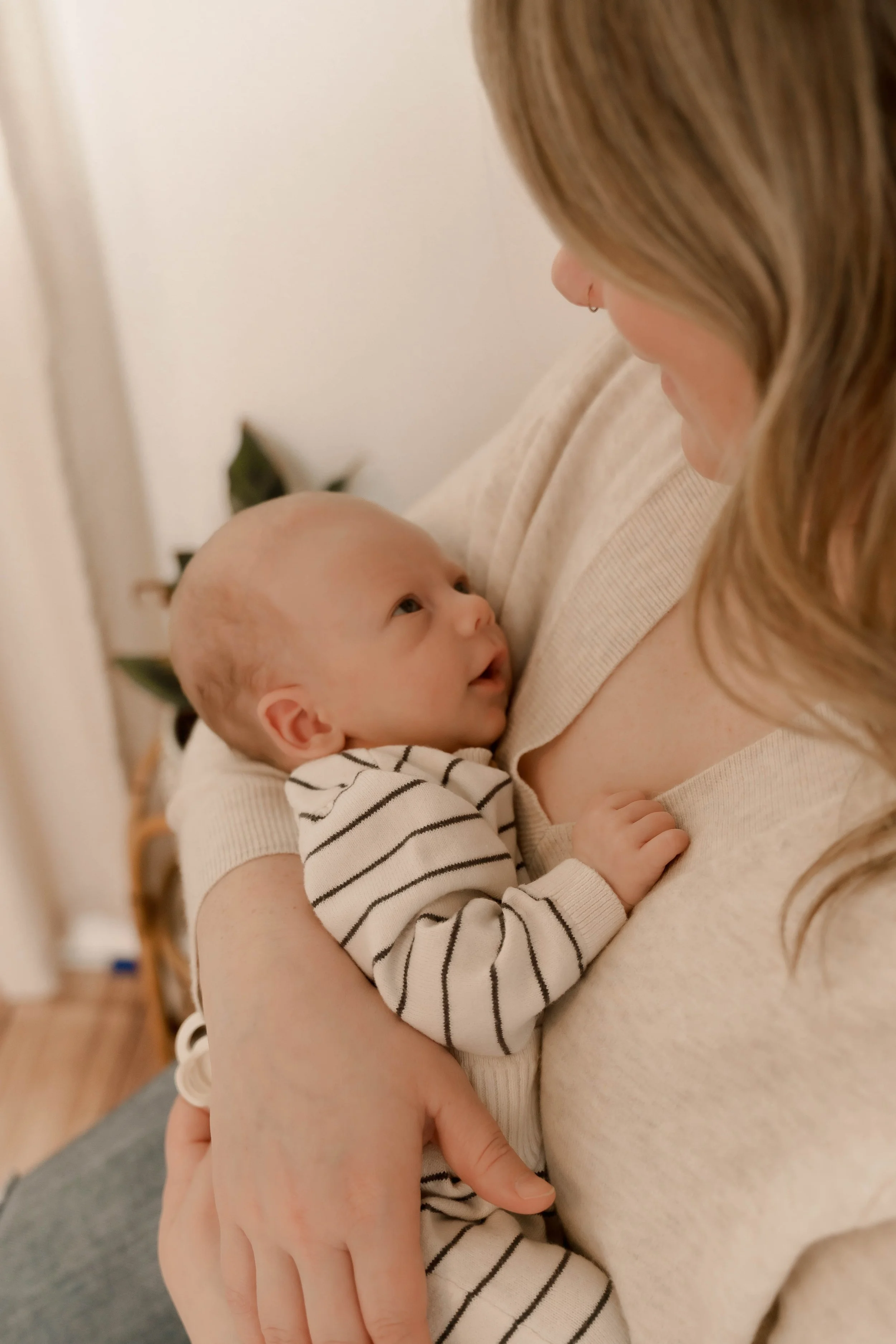 A woman holding an infant in her arms, looking at each other affectionately indoors with a plant and a chair in the background.
