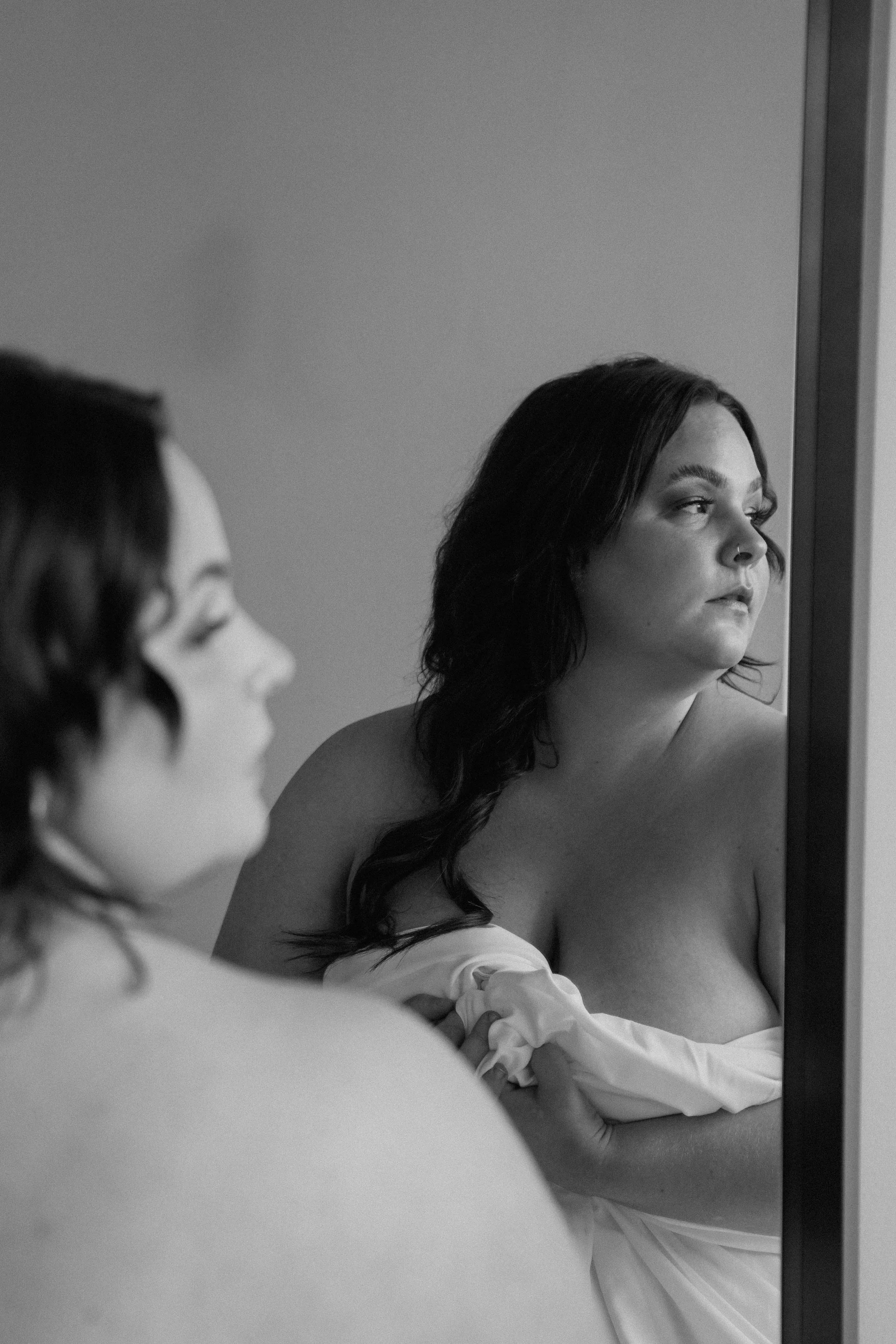 A woman with dark hair looking at herself in a mirror, holding a piece of clothing in her hand.