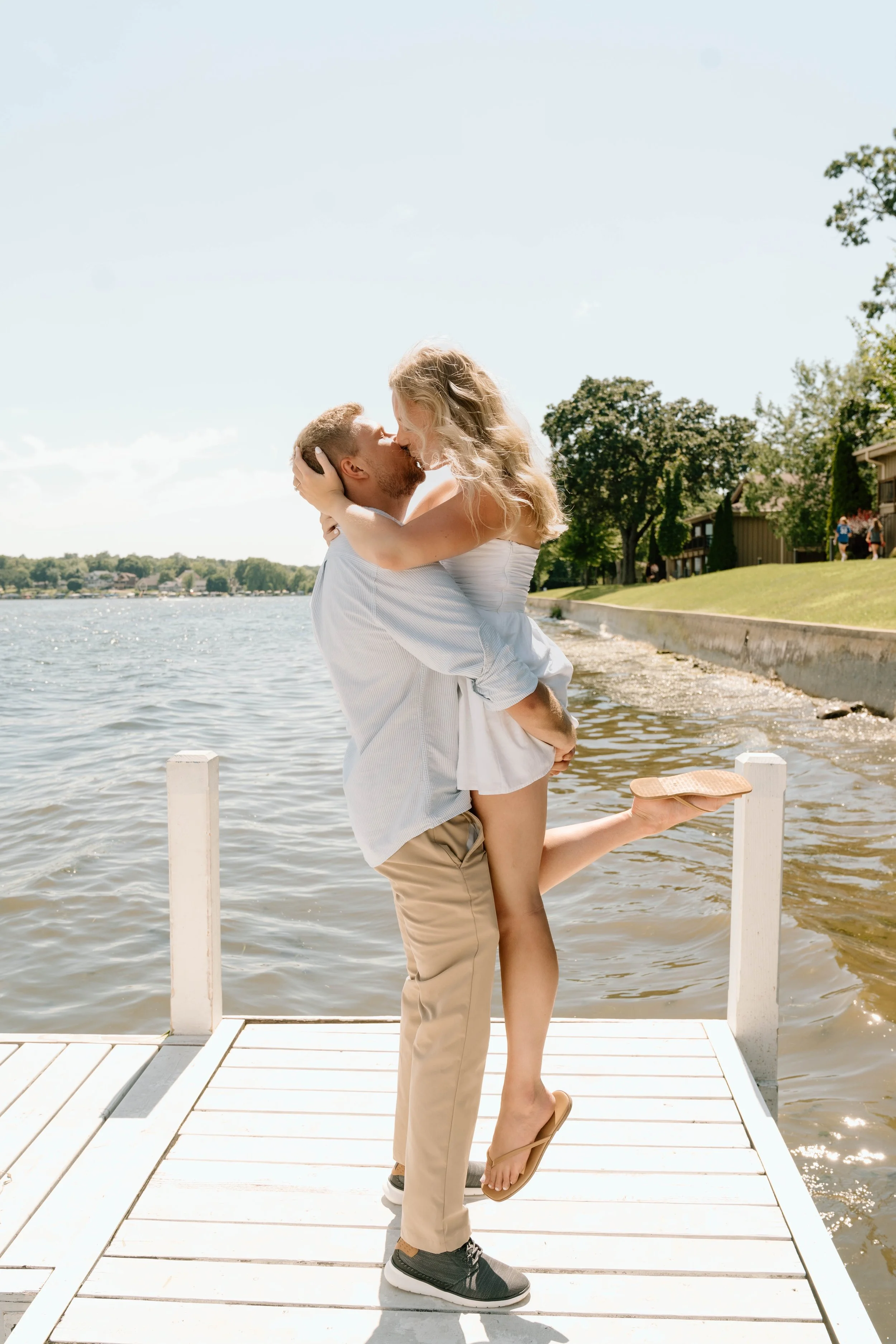 A man lifting a woman in a romantic embrace on a dock by a lake, with trees and houses in the background, under a partly cloudy sky.