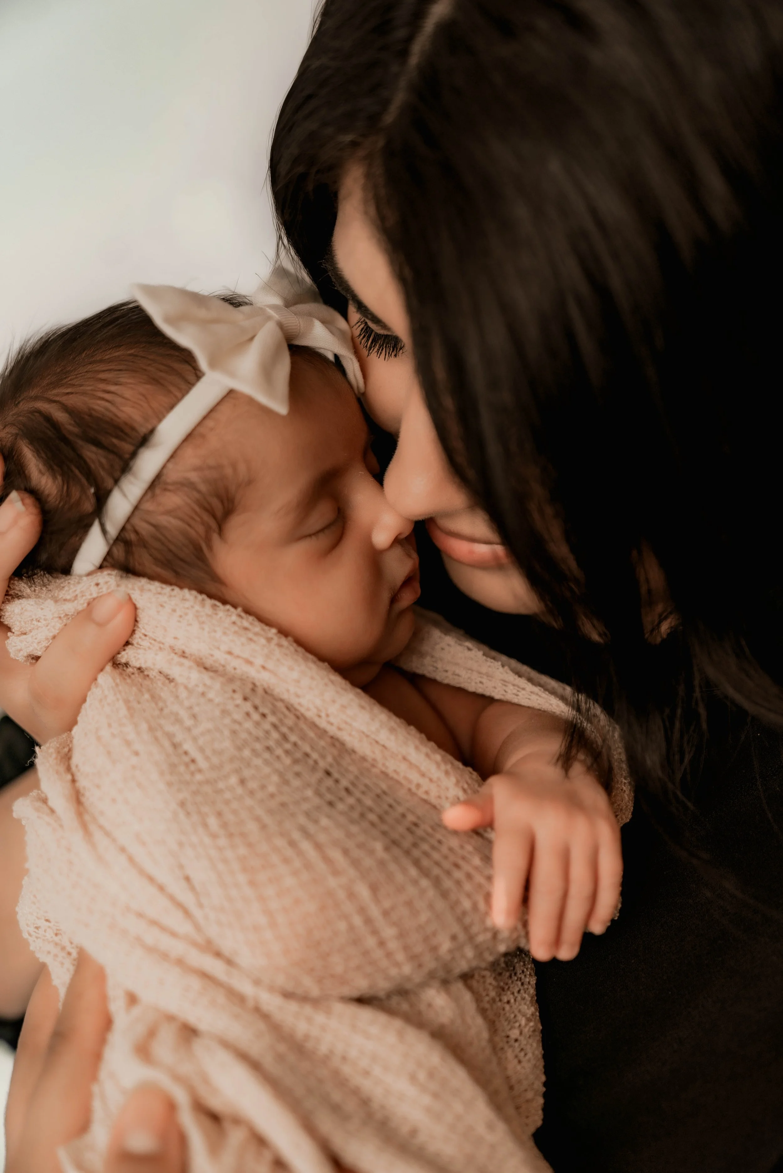A woman with dark hair and long eyelashes gently kisses a sleeping baby girl wrapped in a light brown blanket, holding her close in a tender moment.
