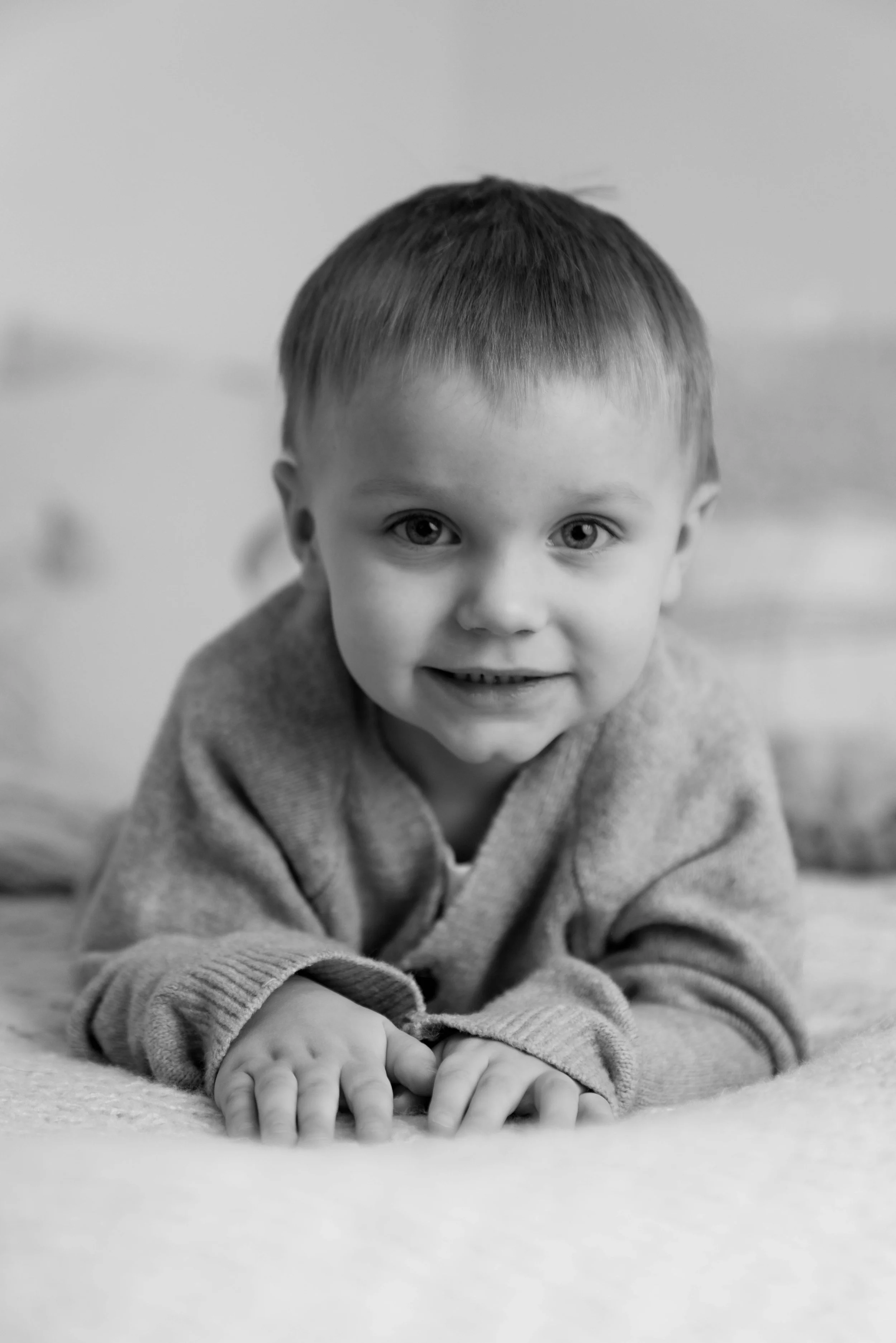 Black and white photo of a young boy lying on his stomach on a bed, smiling at the camera.
