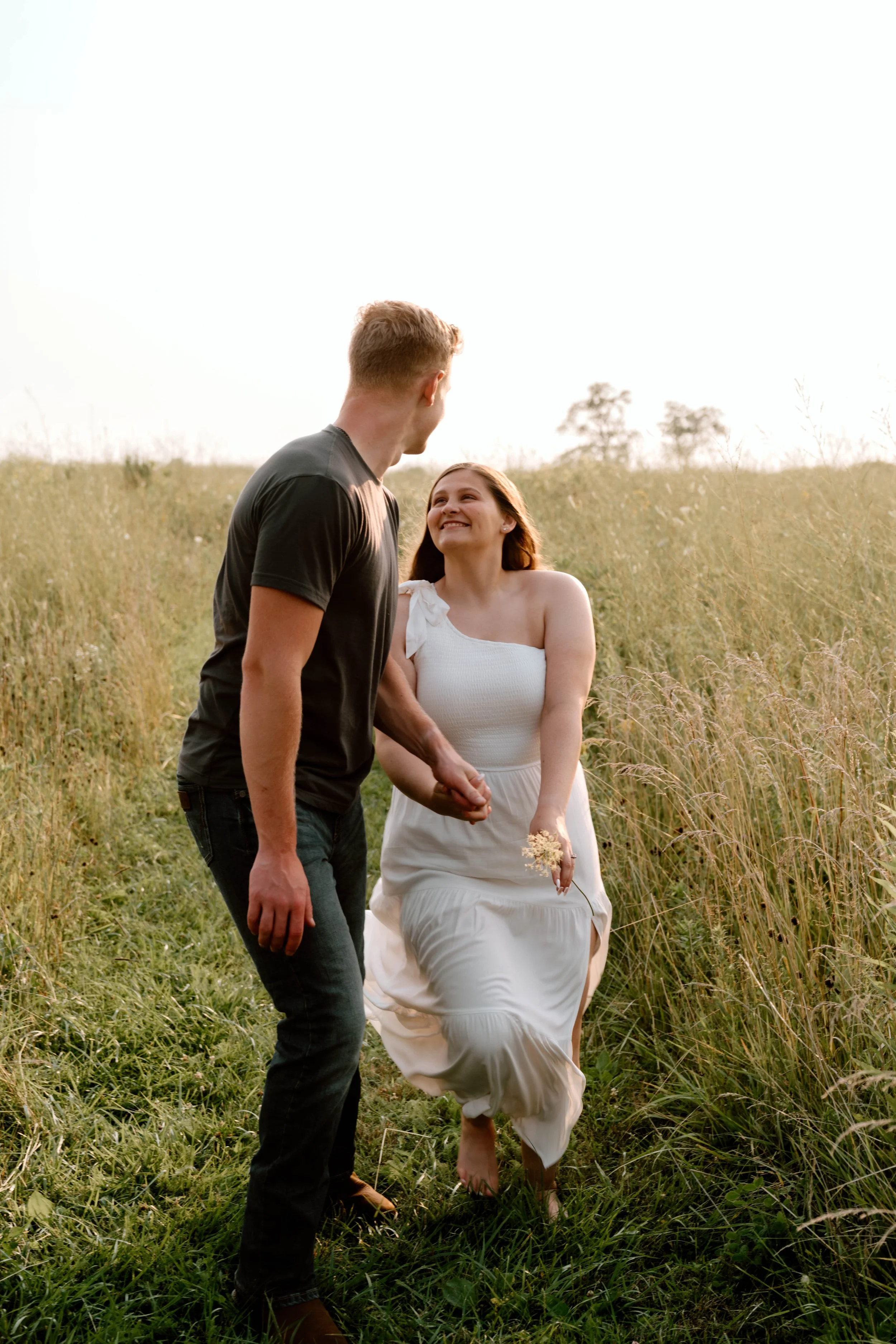 A happy couple holding hands and smiling in a grassy field during sunset, with the woman wearing a white dress and the man wearing a black shirt and jeans.