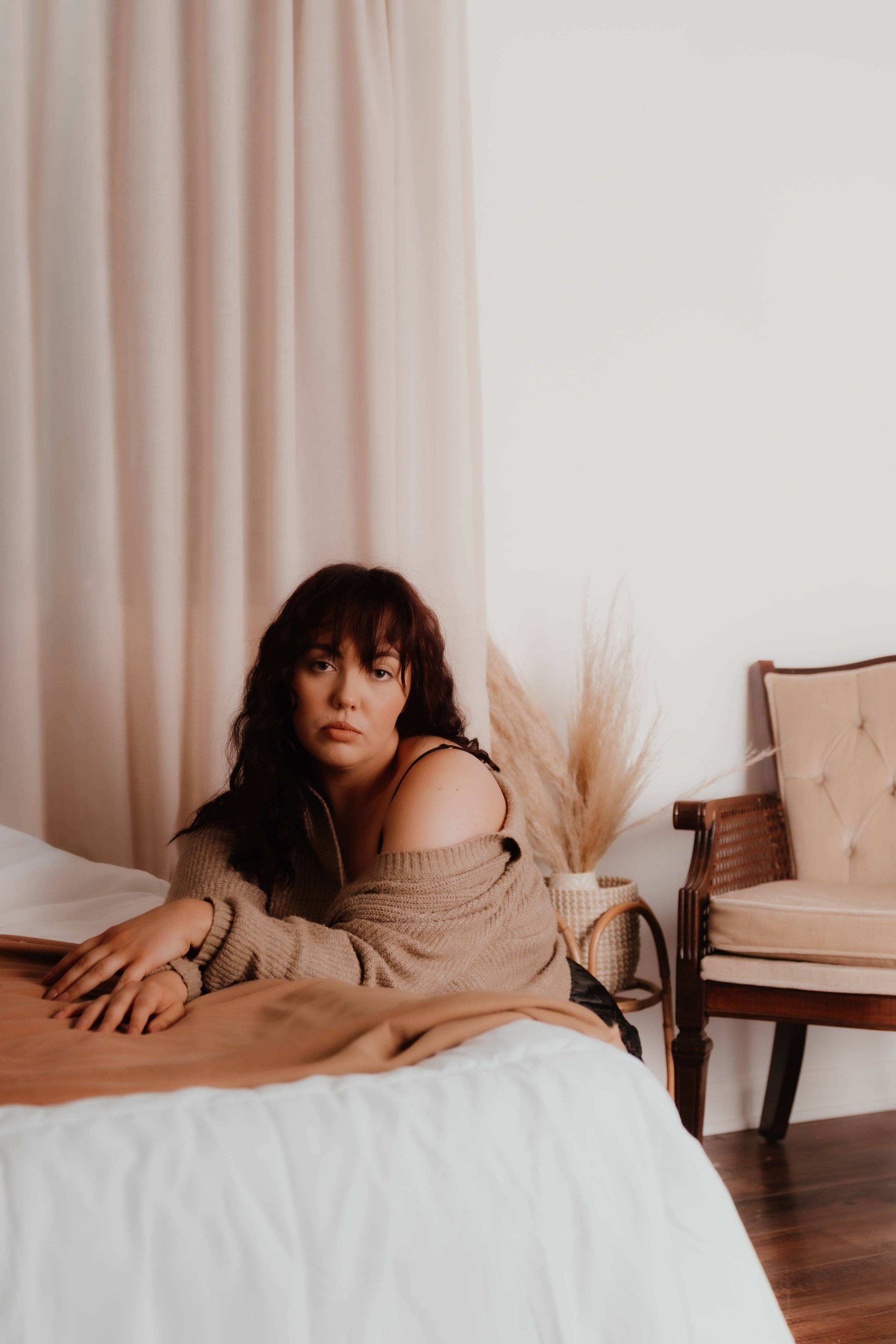 A woman with dark curly hair and light skin lying on her stomach on a bed, wearing a tan sweater that slips off her shoulder, in a softly lit bedroom with neutral decor.
