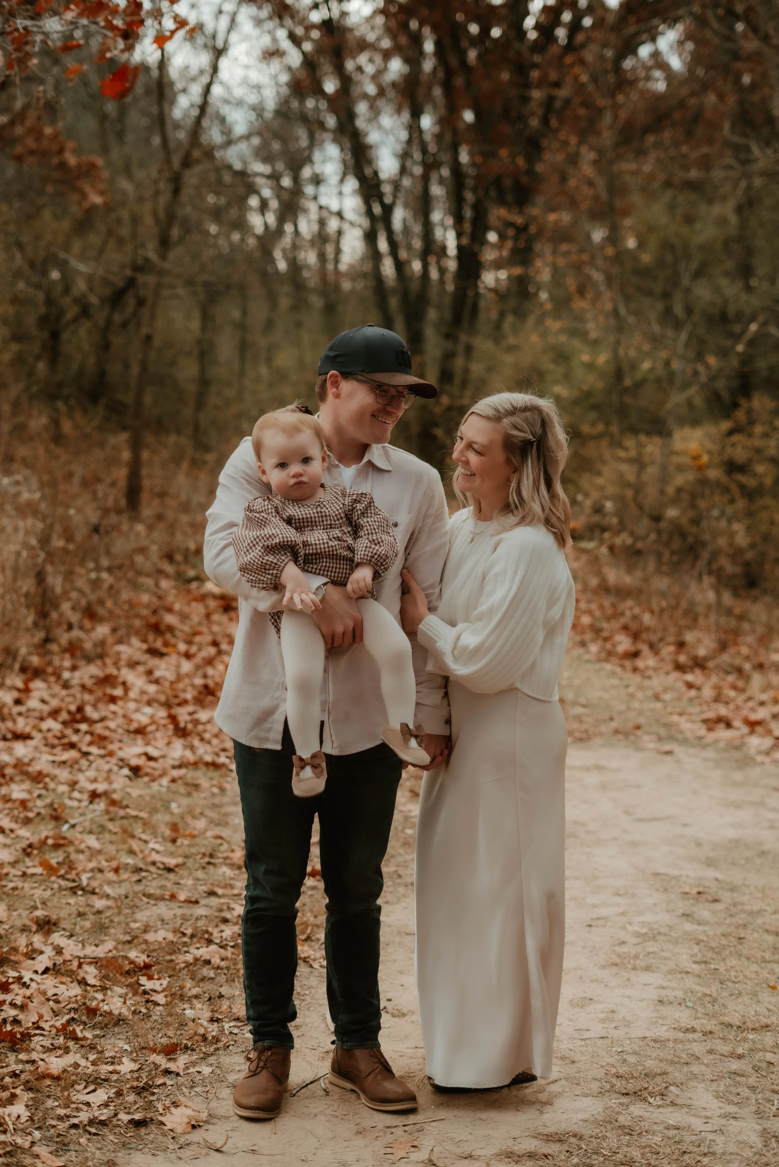 A family of three standing on a wooded trail in autumn, smiling and enjoying each other's company. The man is holding a young child, and the woman is standing next to them, all wearing fall clothing.