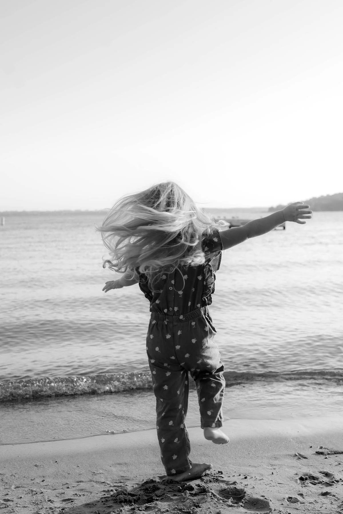 A young girl with long hair running and jumping at the edge of a lake or ocean on a sunny day.