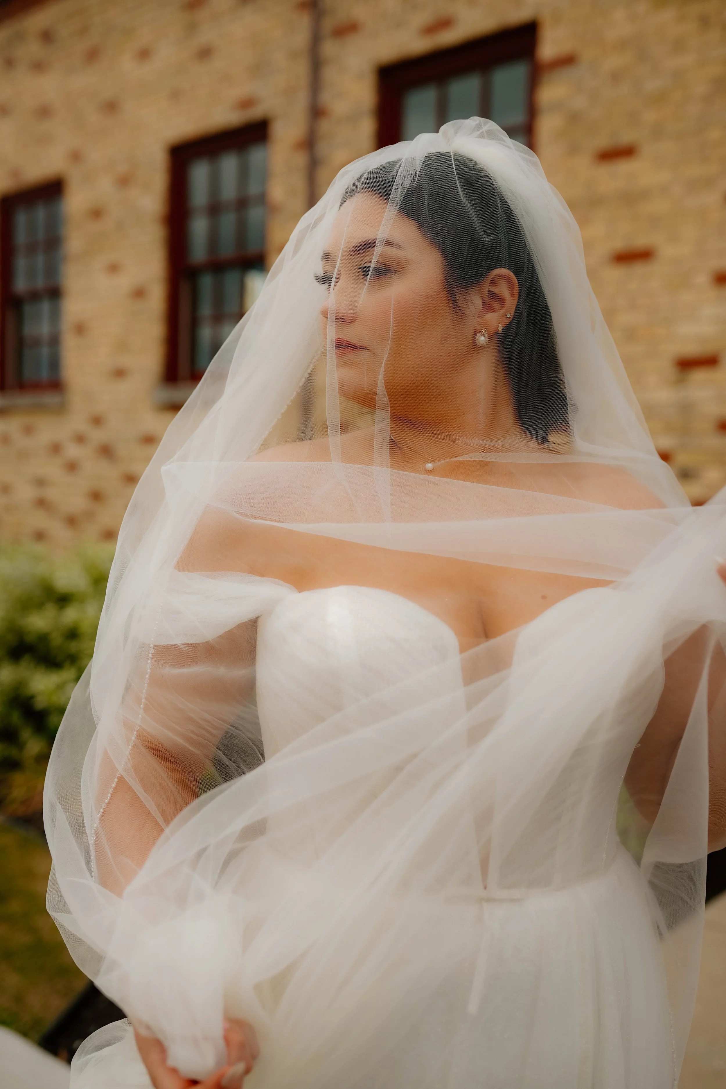 A bride with dark hair and earrings, wearing a wedding dress and veil, standing outdoors in front of a brick building with multiple windows.