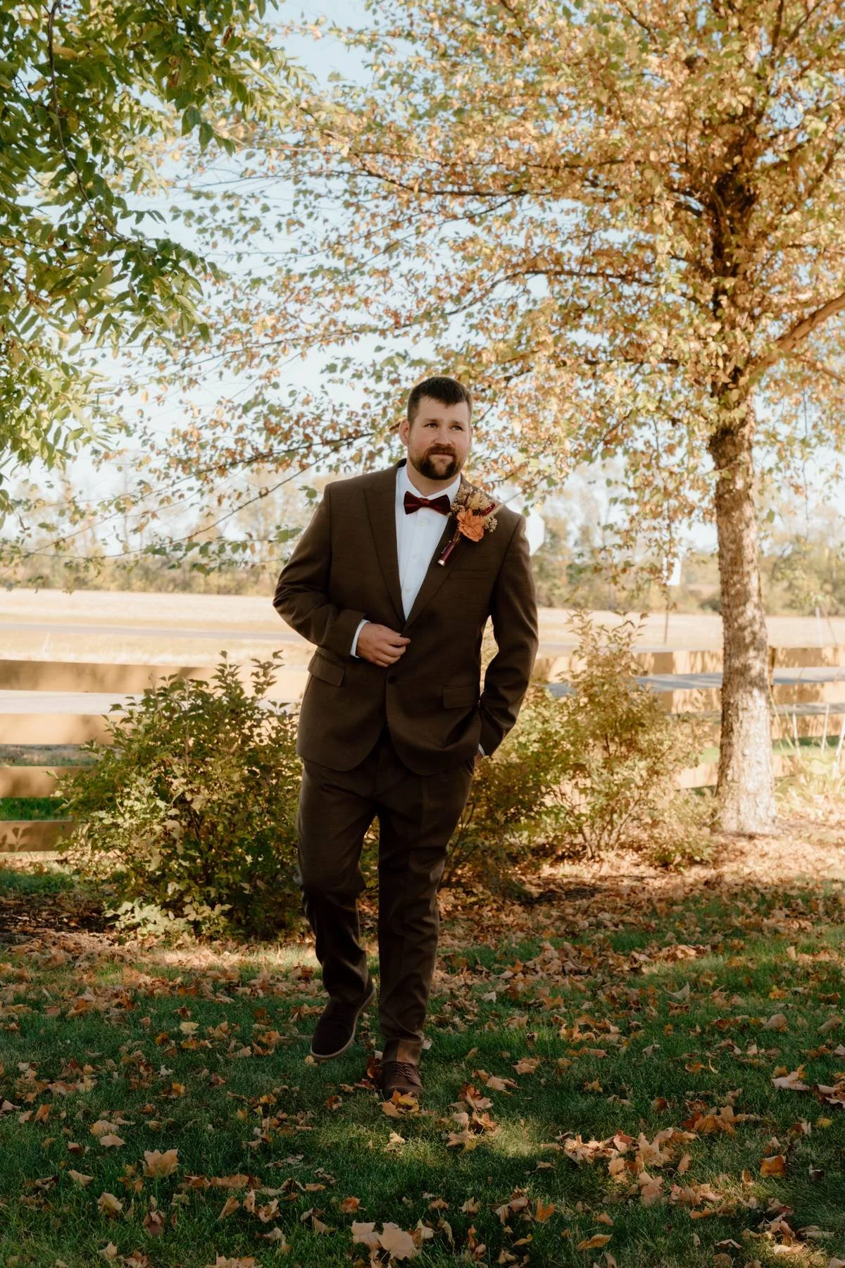 Man in a brown suit and red bow tie standing outdoors on grass, with trees and a wooden fence in autumn foliage background.
