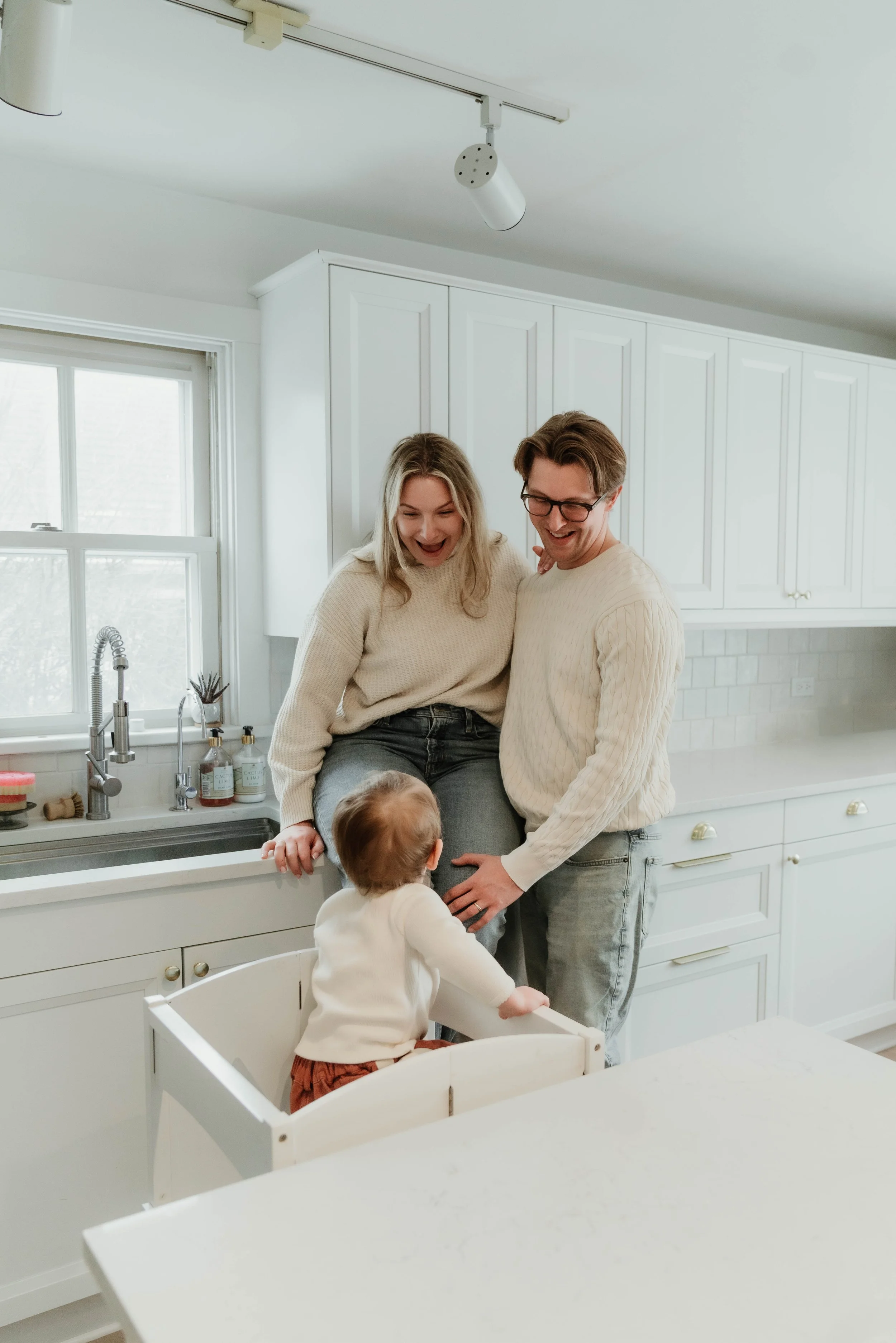 A family of three, including a woman, man, and young child, happily interacting in a bright, white kitchen.