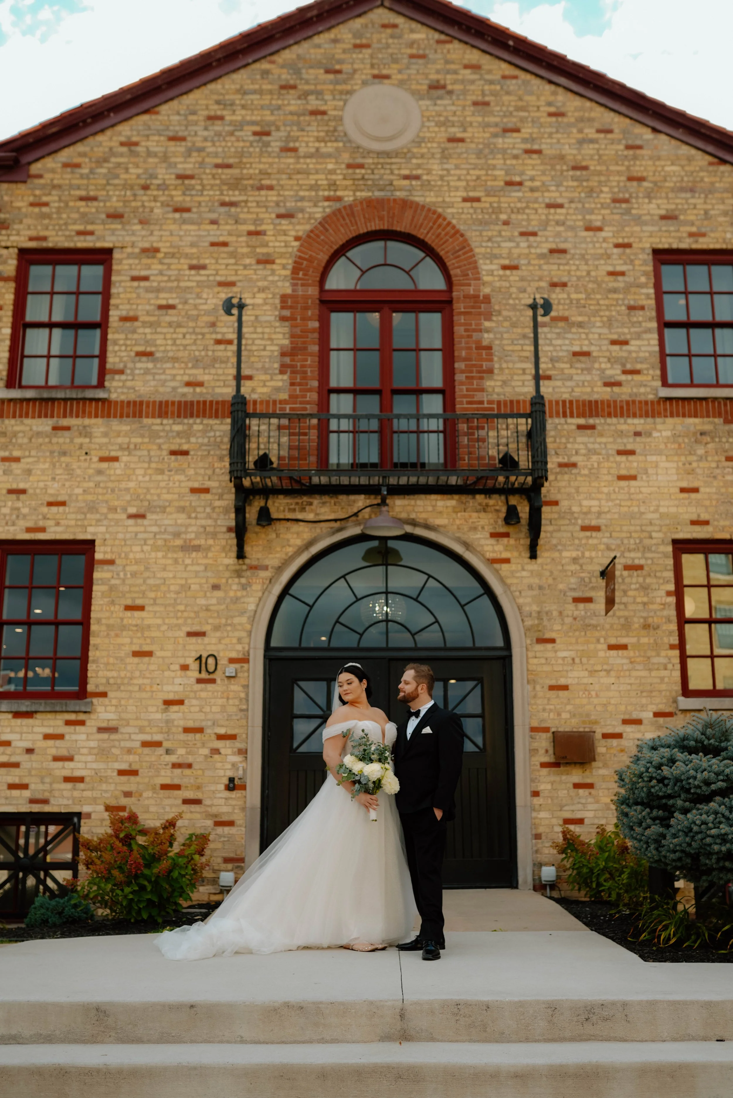 A bride in a white wedding gown and a groom in a black tuxedo standing outside a brick building with large windows, a balcony, and an arched entrance. The bride is holding a bouquet of flowers, and they are looking at each other.