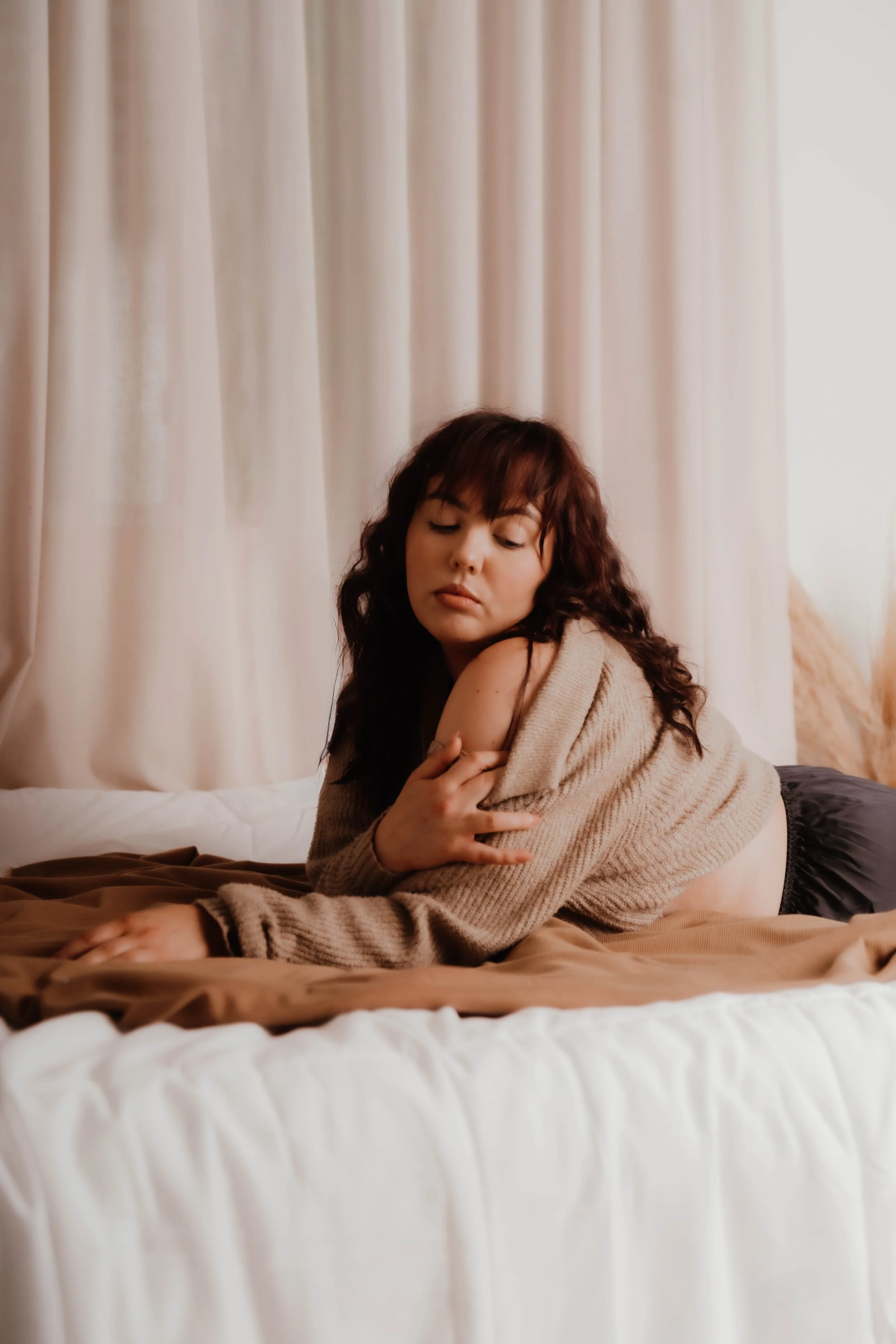 A woman with curly dark hair sitting on a bed, wrapped in a beige sweater, looking down with a contemplative expression, in a softly lit room with cream-colored curtains.