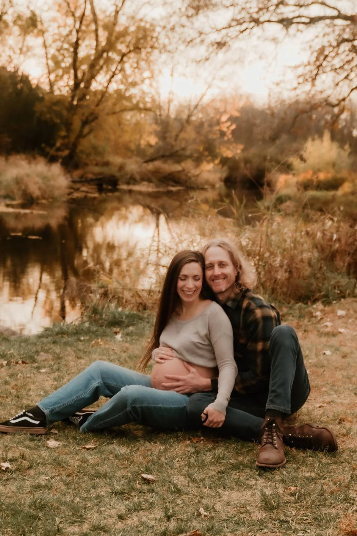A pregnant woman and a man sitting on the grass near a lake, smiling and embracing, during sunset with autumn trees in the background.