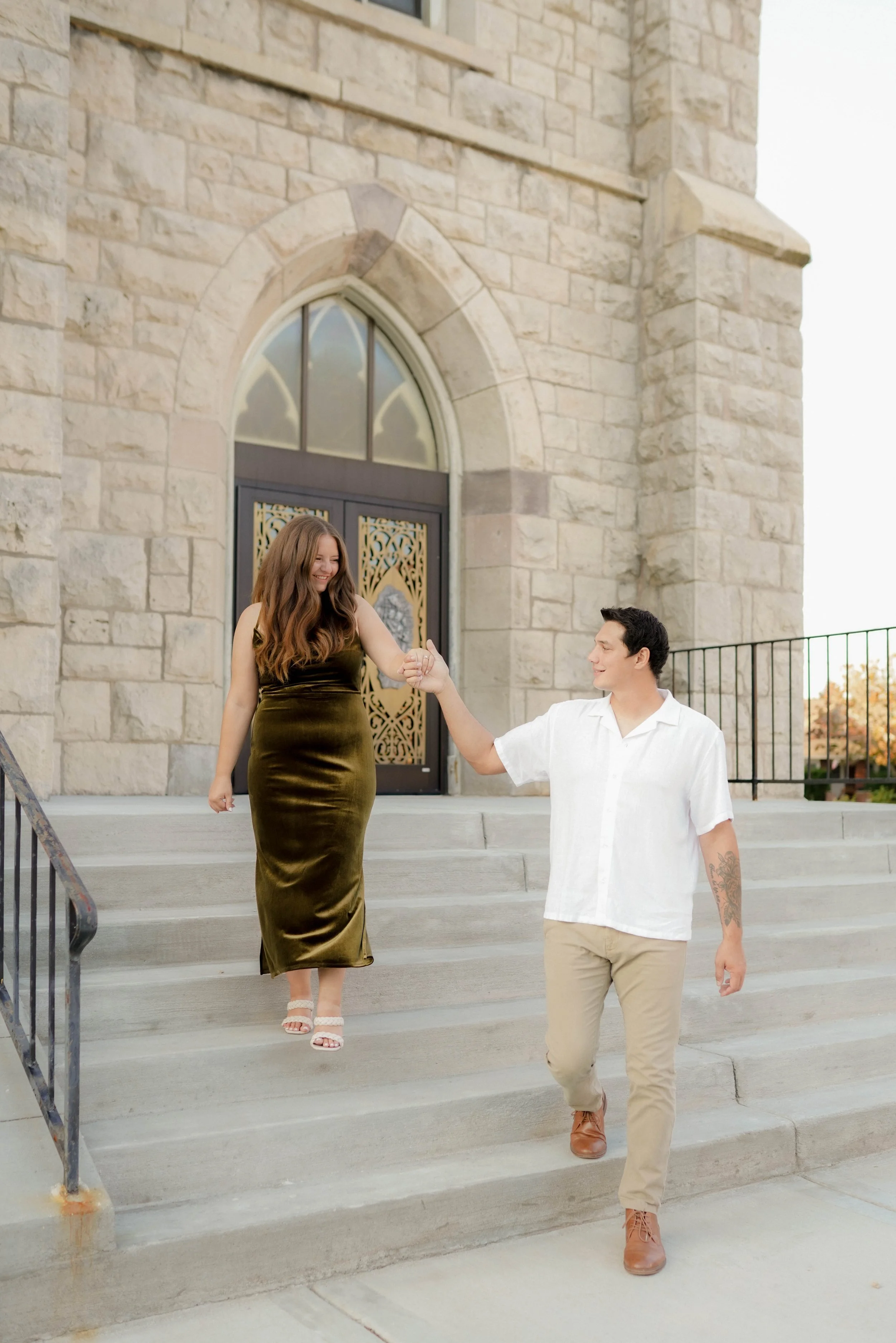 A man and a woman are holding hands and walking down the stairs outside a stone building with arched windows and a decorative door, smiling at each other.