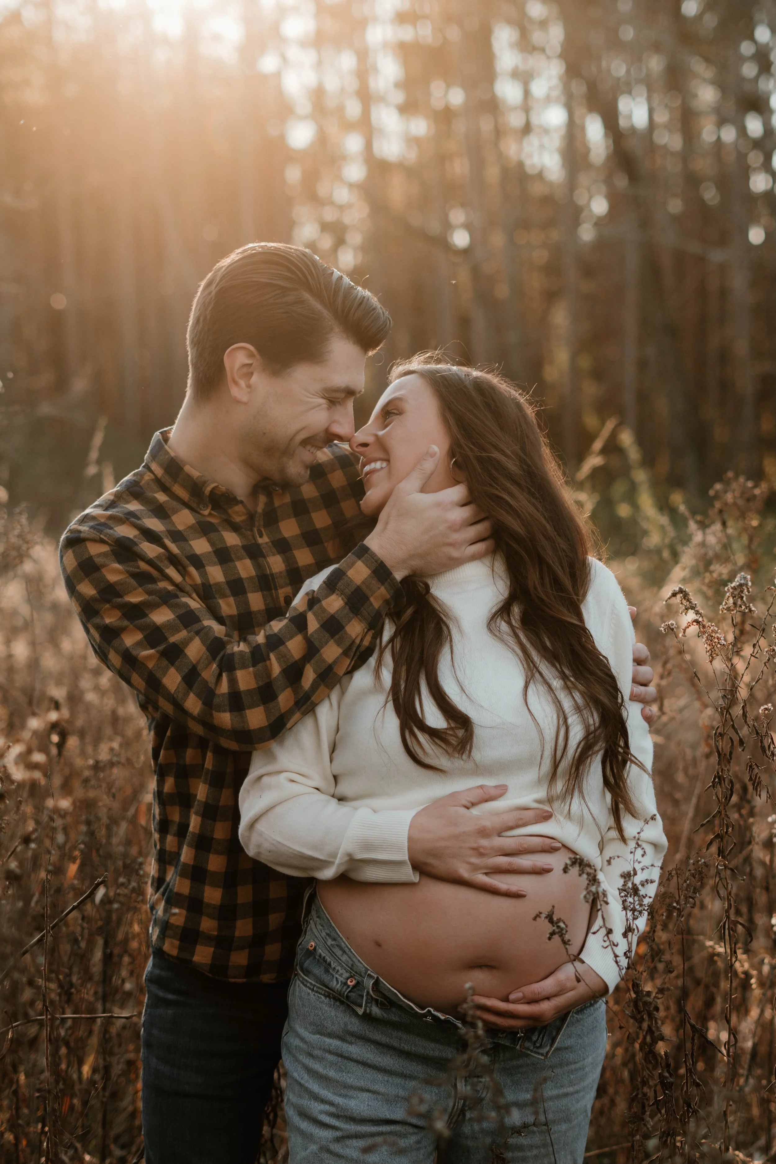 A pregnant woman and a man are smiling and touching foreheads in a field with autumn foliage, bathed in warm sunlight.