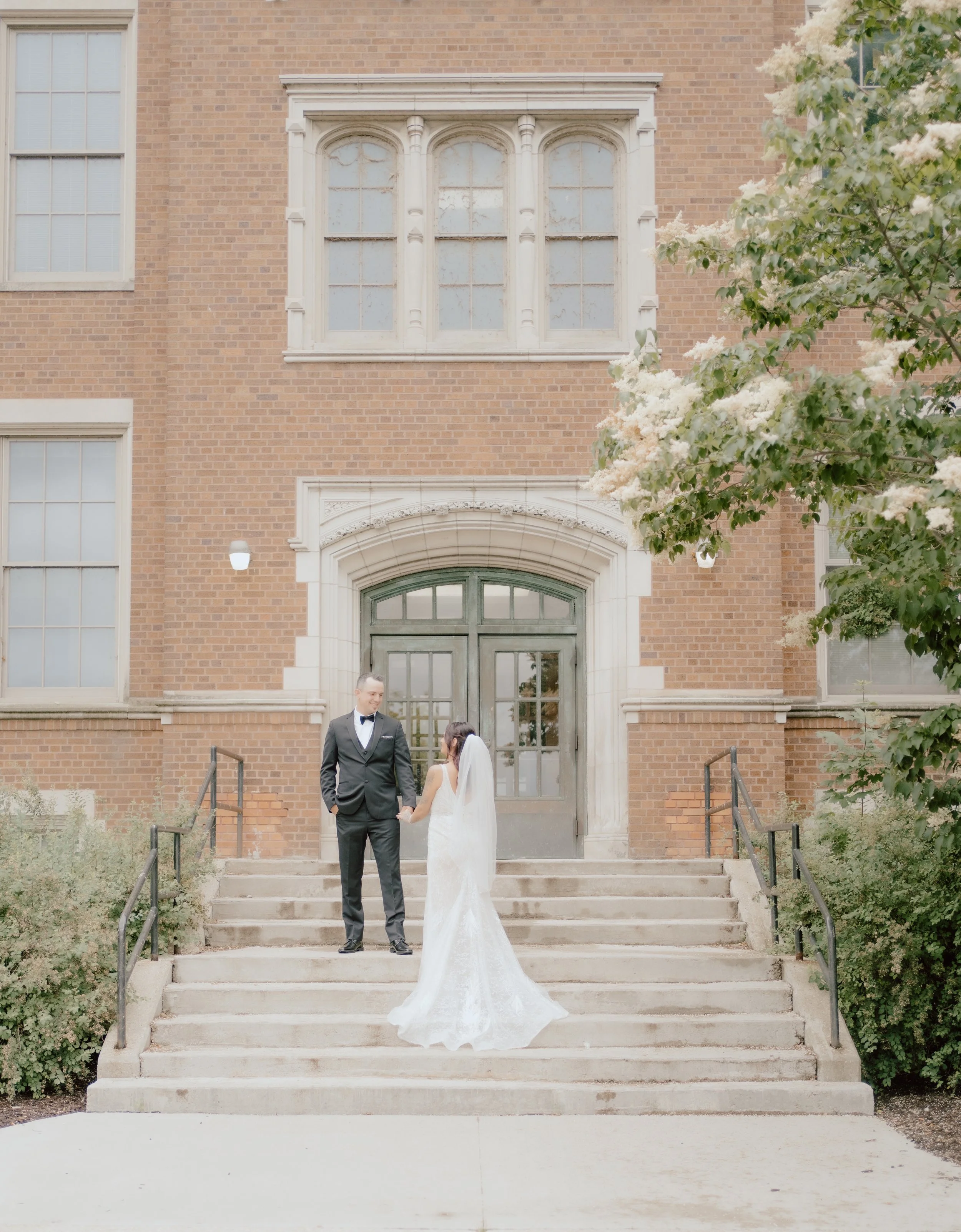 A bride and groom holding hands and looking at each other on a staircase outside a brick building during a wedding or engagement photo shoot. The bride is wearing a white wedding dress and veil, and the groom is in a black tuxedo with a bow tie. Ther