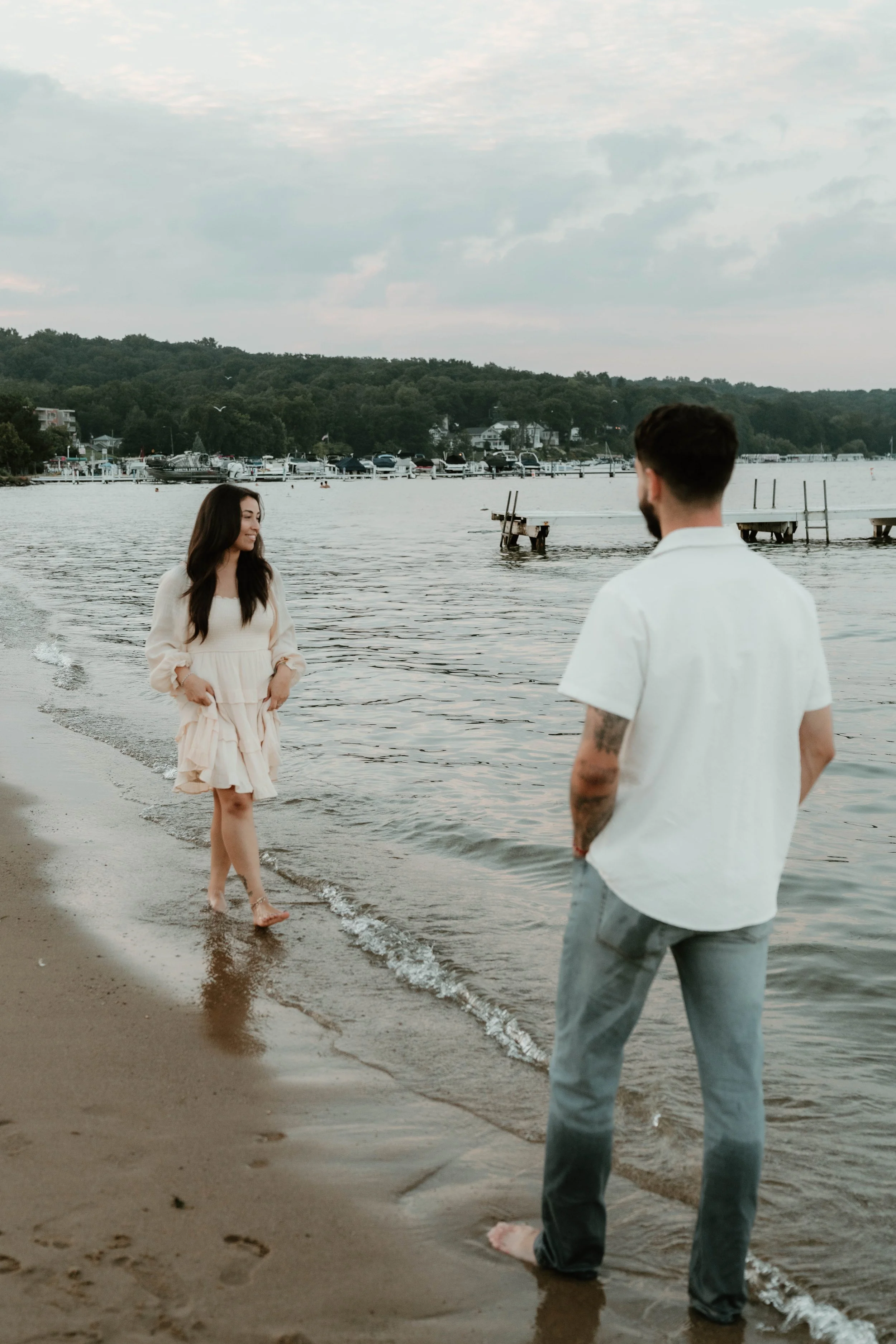 A woman in a light-colored dress walking barefoot along the beach, smiling at a man standing in the water near the shore, with a harbor and boats in the background.