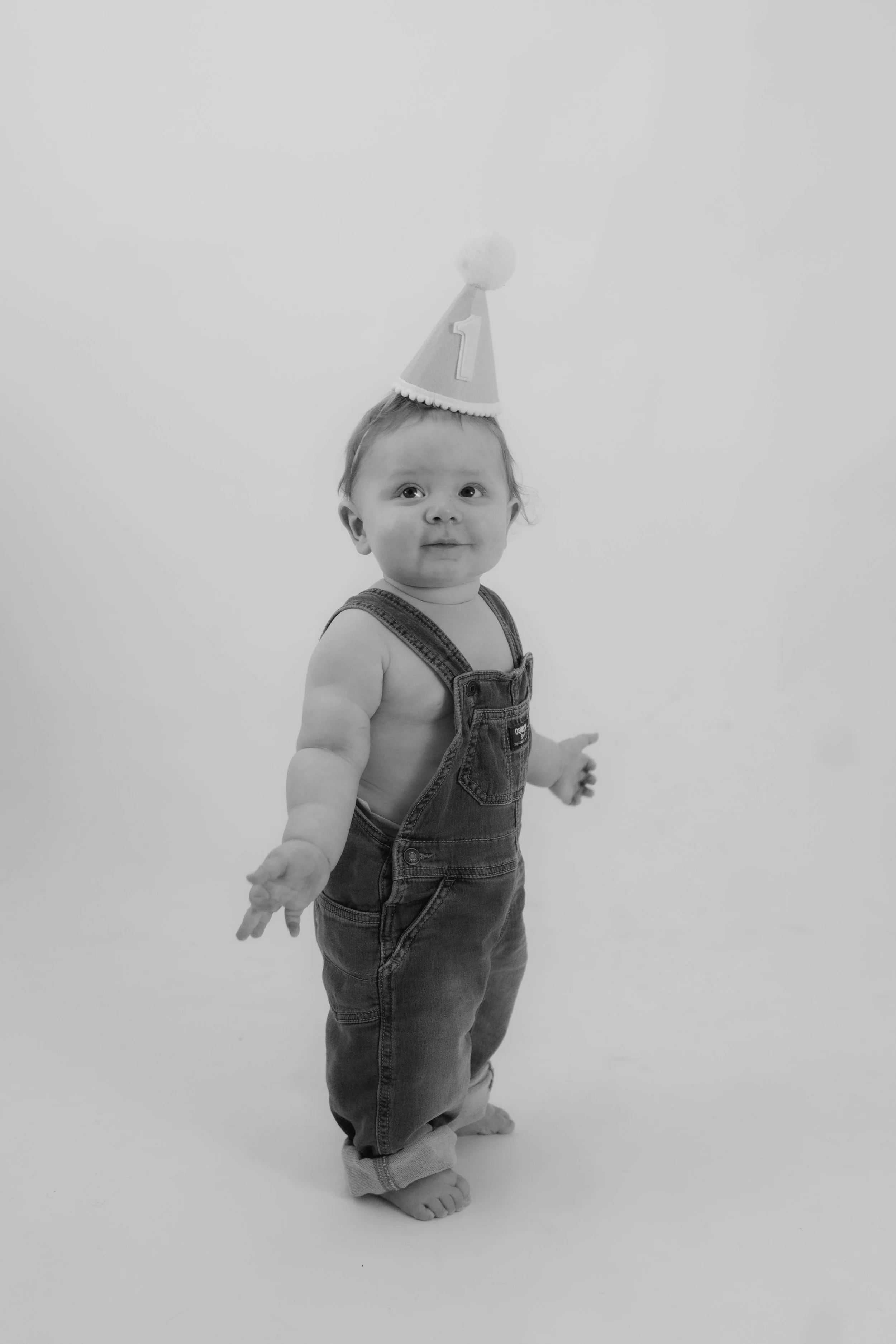 Baby wearing overalls and a birthday hat with the number one, standing barefoot on a plain background.