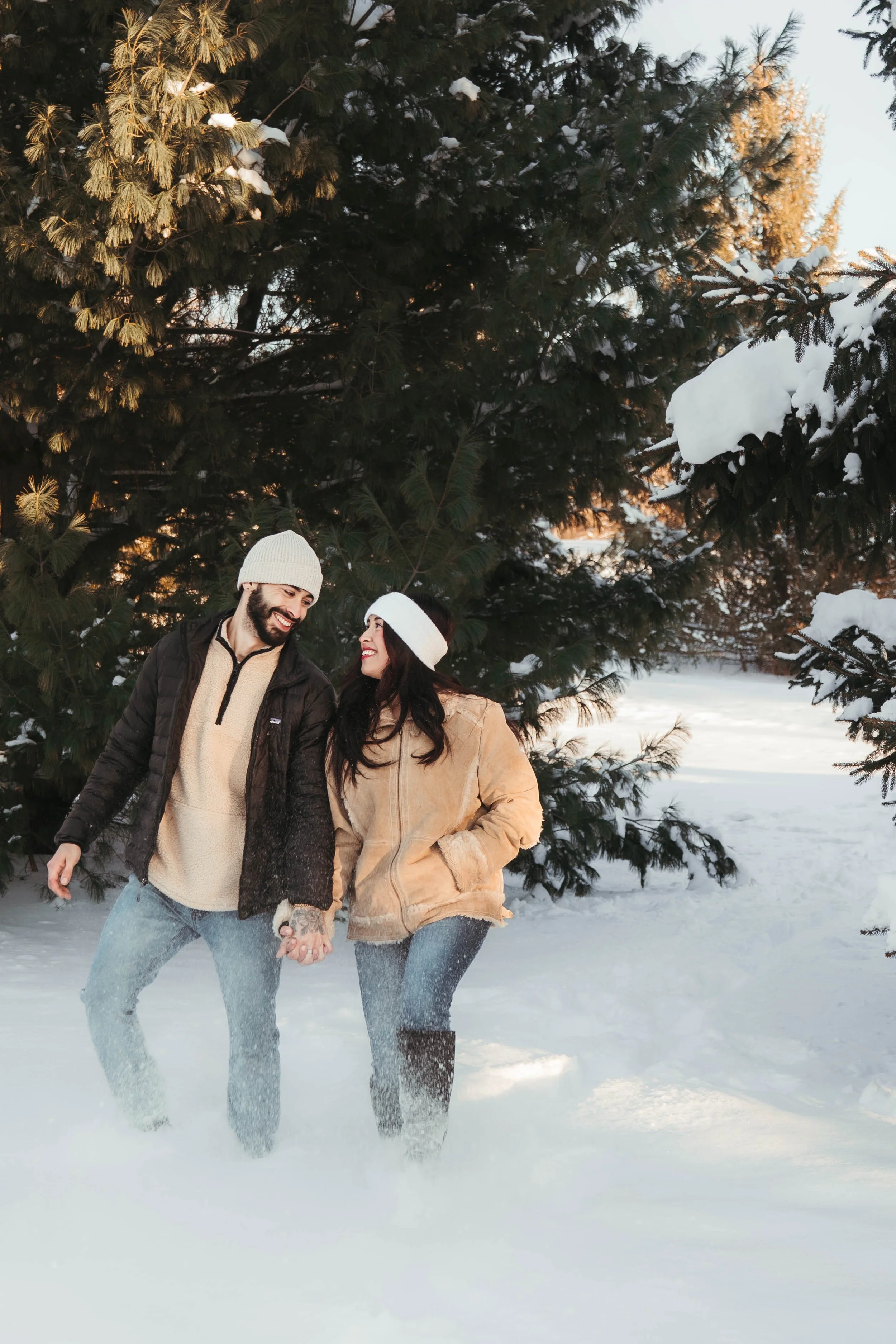 A smiling man and woman holding hands in a snowy landscape, standing in front of evergreen trees with snow on their branches, during sunset.