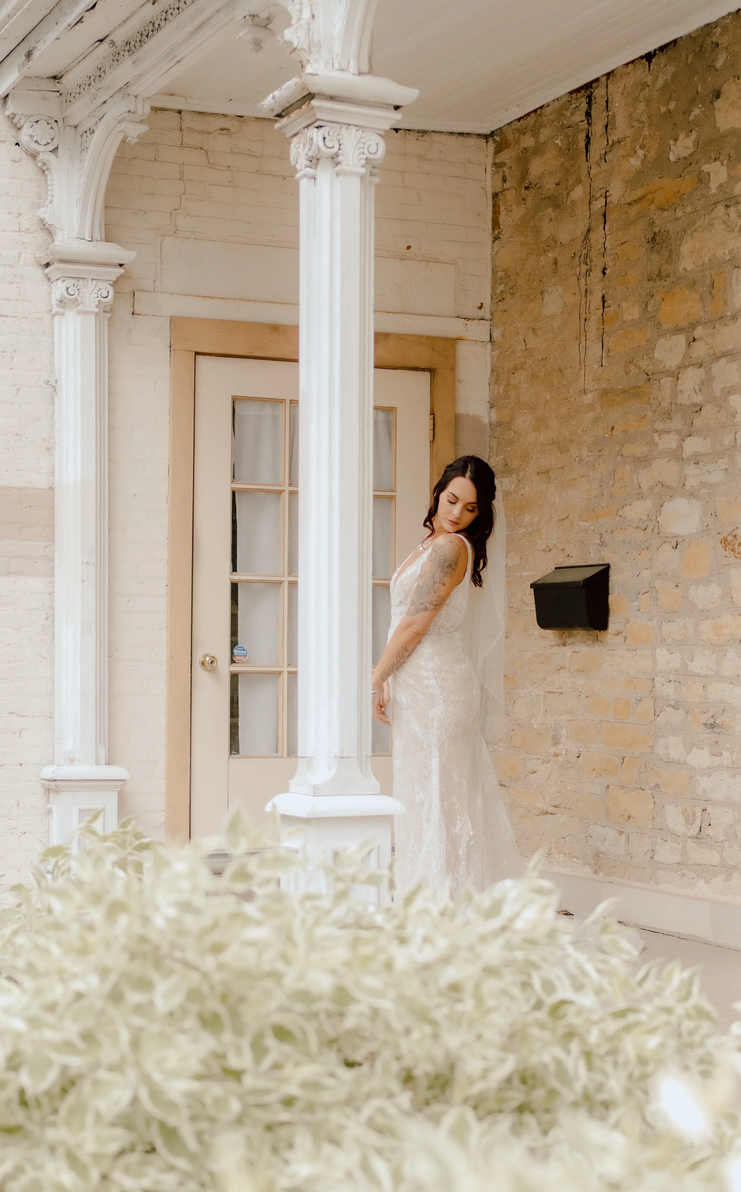 A woman in a white lace wedding dress stands indoors near a brick wall and white door, partially obscured by a large bouquet of white flowers in the foreground.