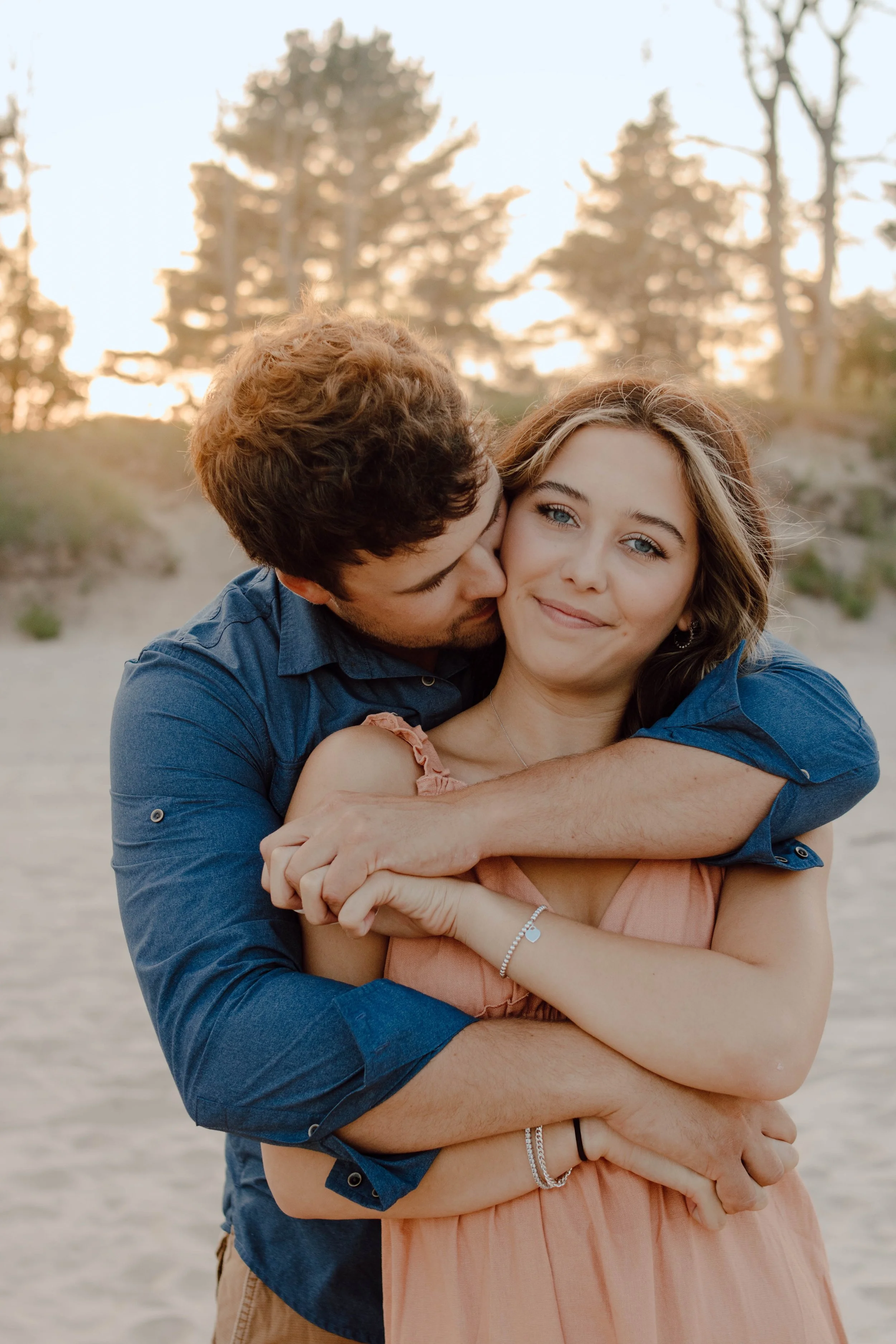A happy couple embracing each other on a sandy beach with trees in the background during sunset.