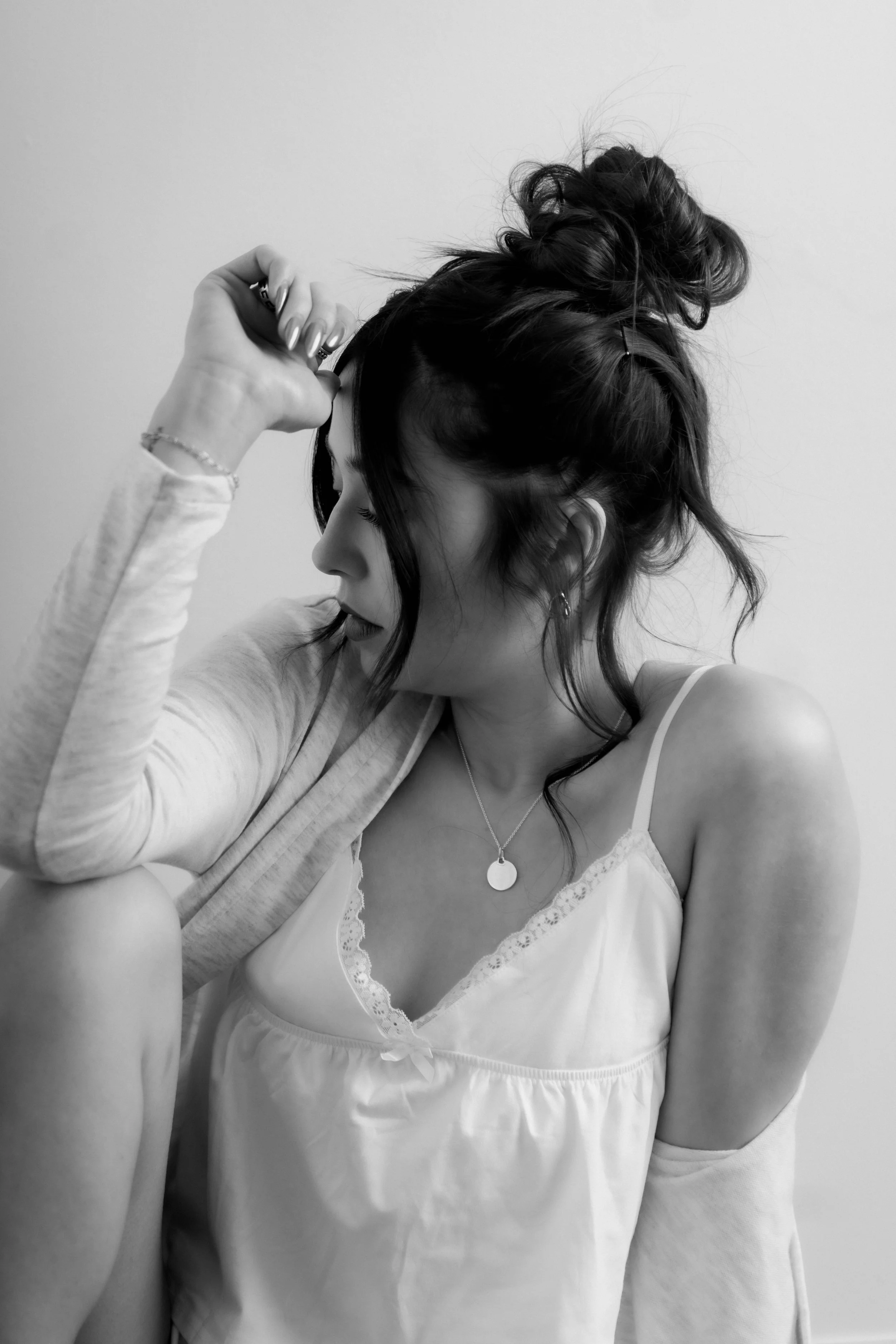 A woman with dark hair in a messy bun, wearing a spaghetti strap top and a necklace, is sitting with her PR arm raised to her head, looking down with a contemplative expression.
