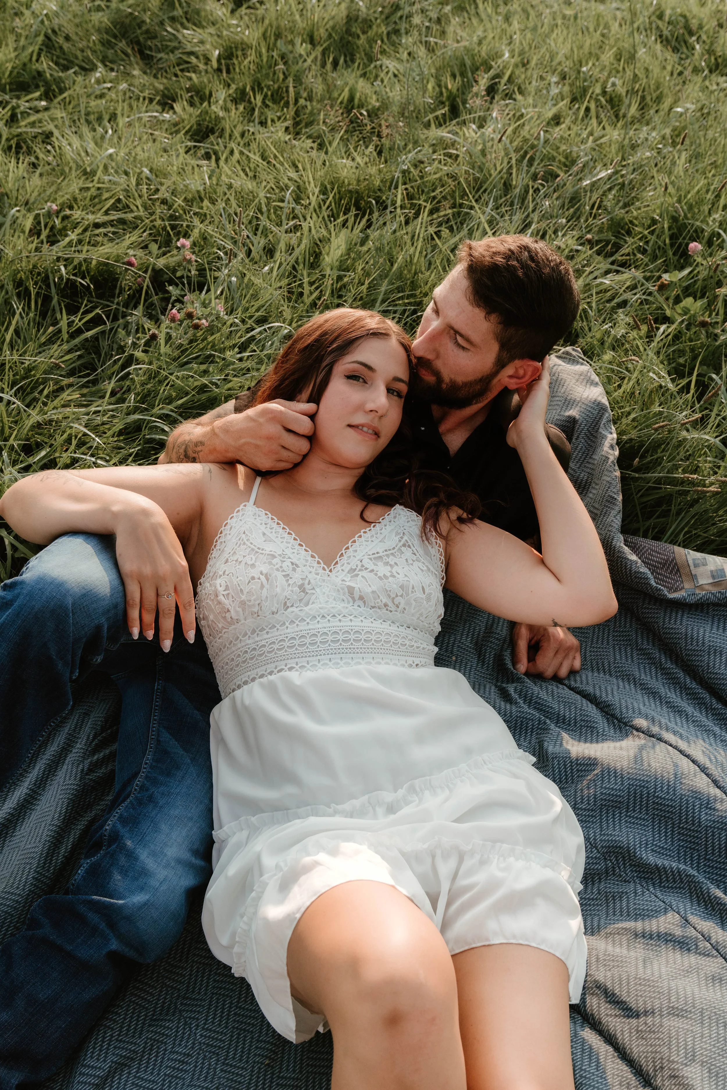 A couple lying on a blanket in a grassy field, with the man gently holding the woman's face as they gaze at each other.