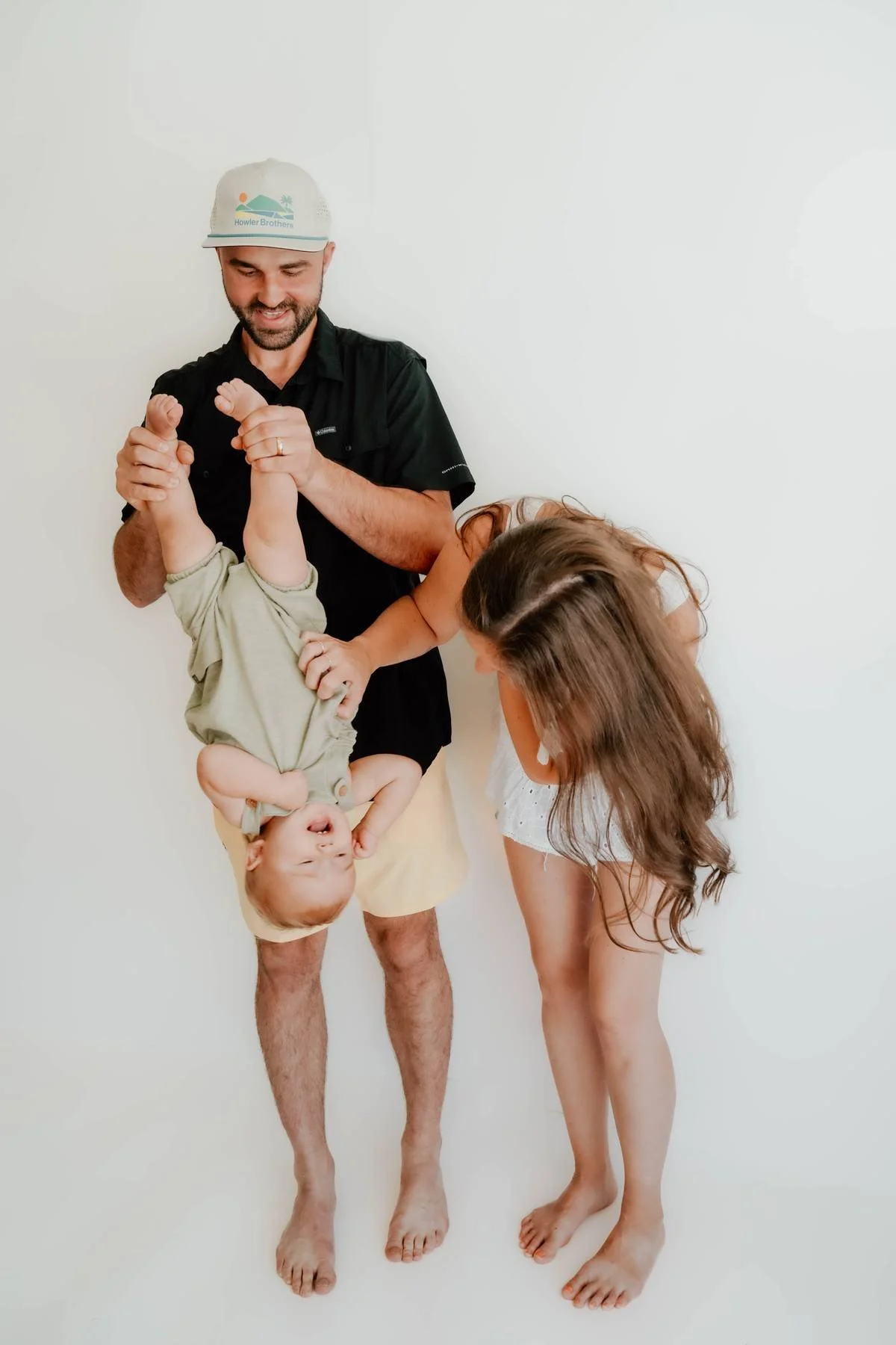 Family of three, a man, woman, and baby, in a playful upside-down pose against a plain white wall. The man is holding the baby upside down by the legs, smiling, while the woman, bending over, touches the baby's foot, both appearing to enjoy the momen