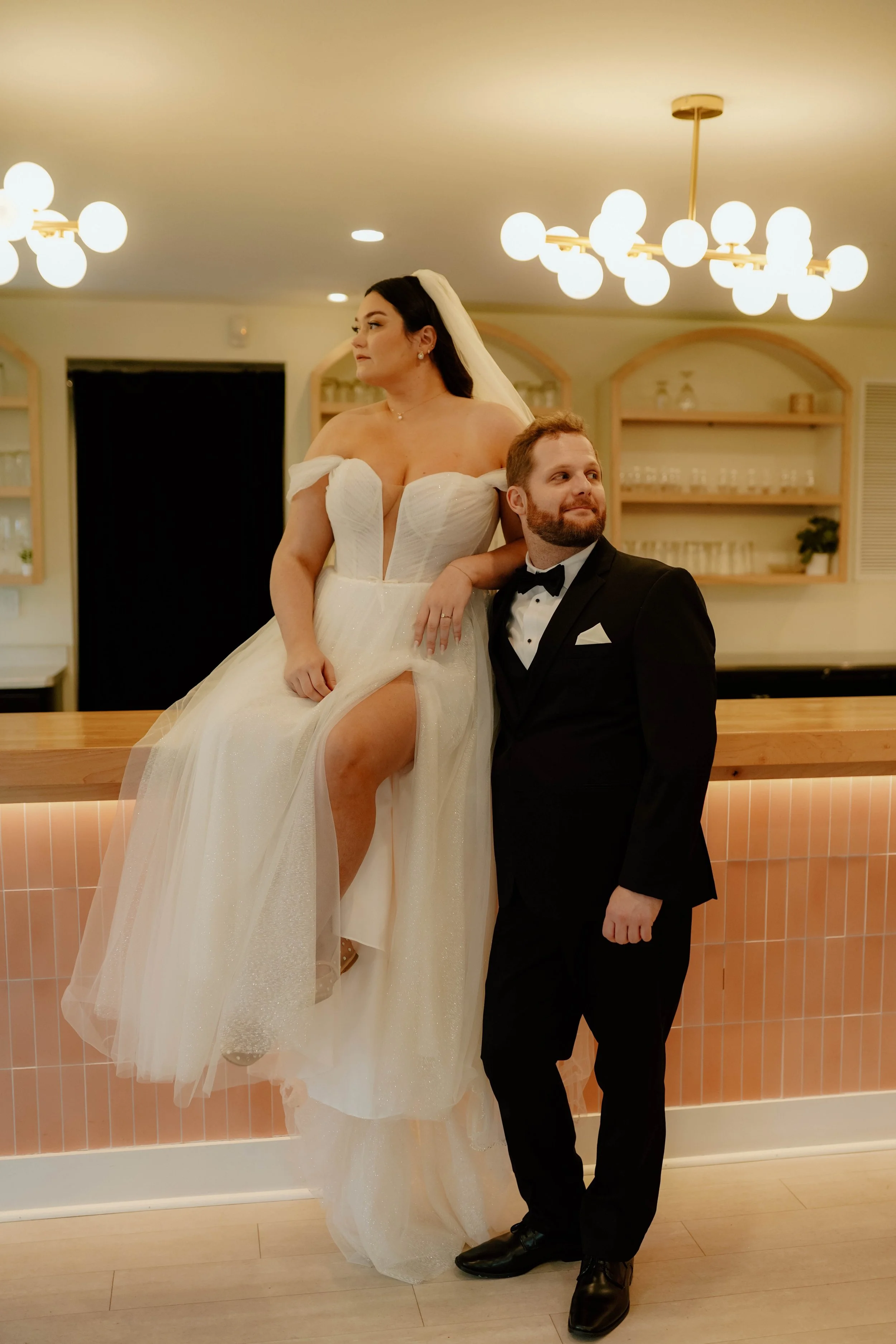 A bride in a white wedding dress with off-the-shoulder sleeves sitting on a counter, and a groom in a black tuxedo standing beside her in an indoor setting with warm lighting and modern decor.