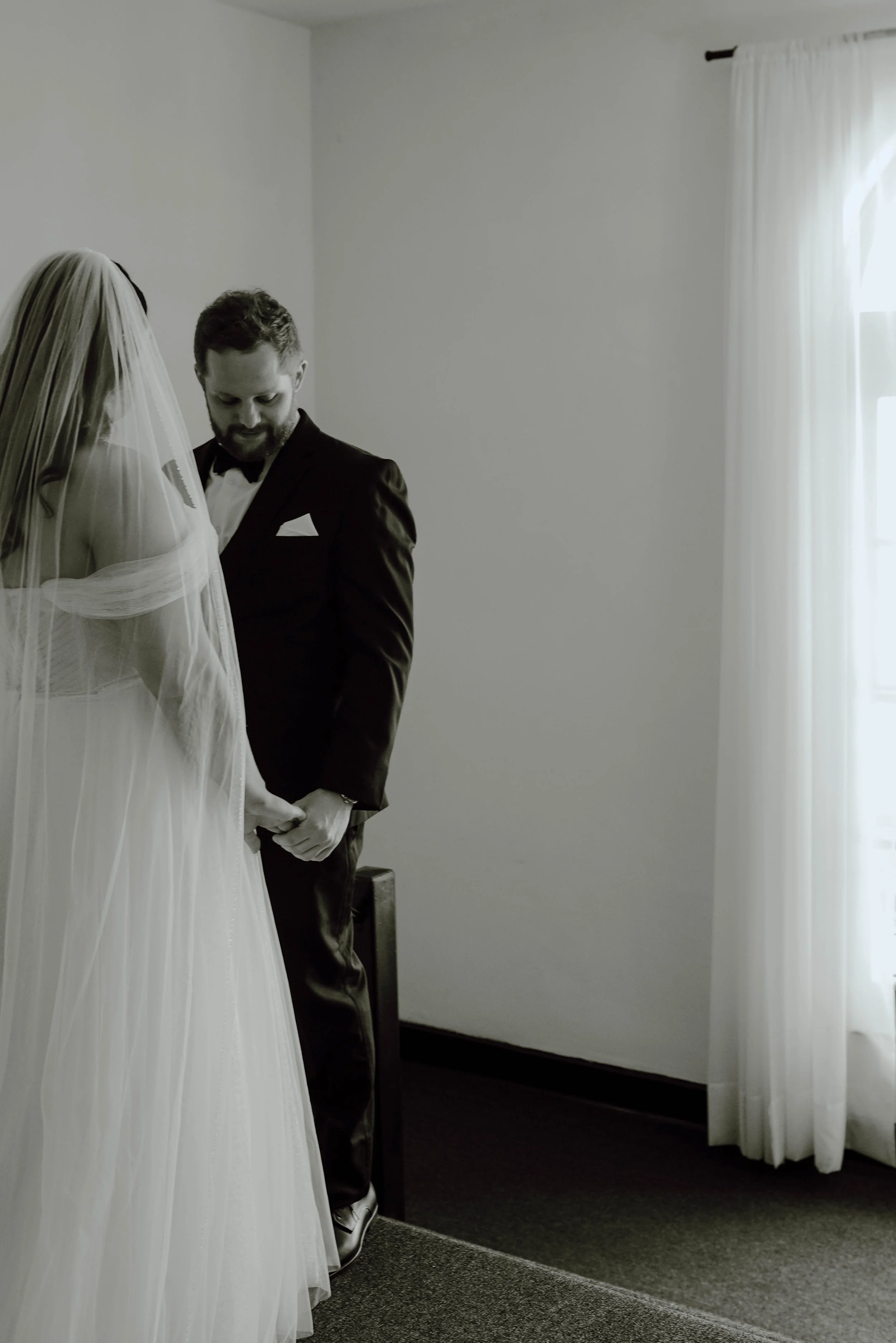 A bride and groom standing close together in a room with natural light, the bride in a white wedding dress with a veil, the groom in a black tuxedo.