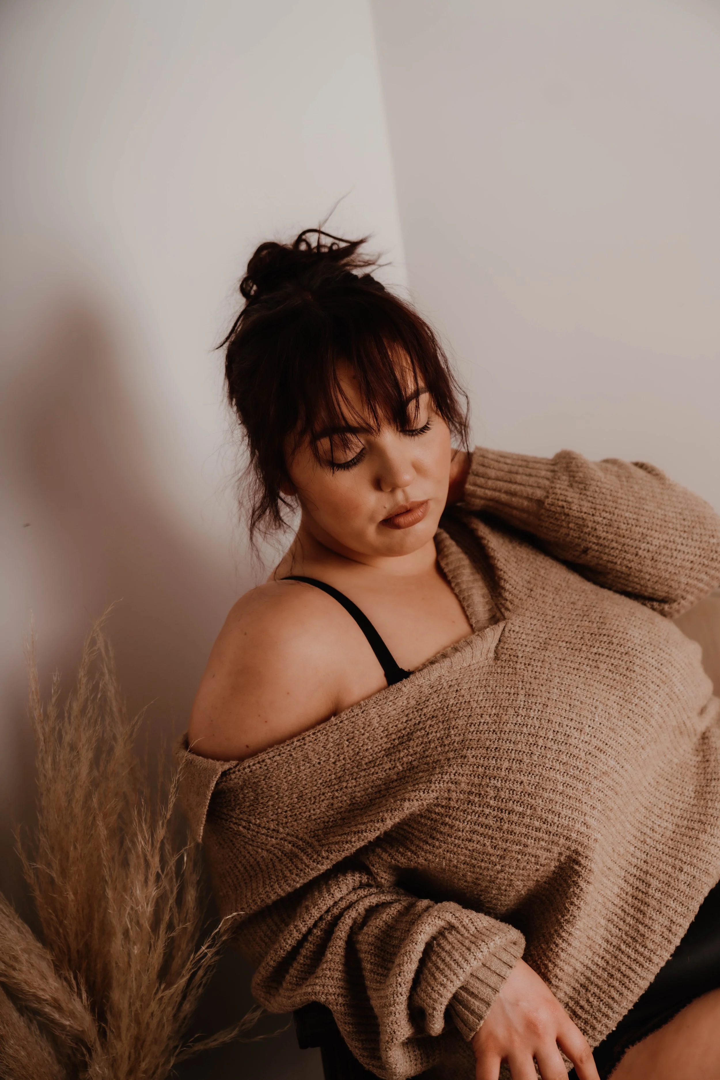 A woman with dark hair styled in a messy bun, wearing a beige sweater off one shoulder and a black strap top, leaning against a wall with closed eyes.