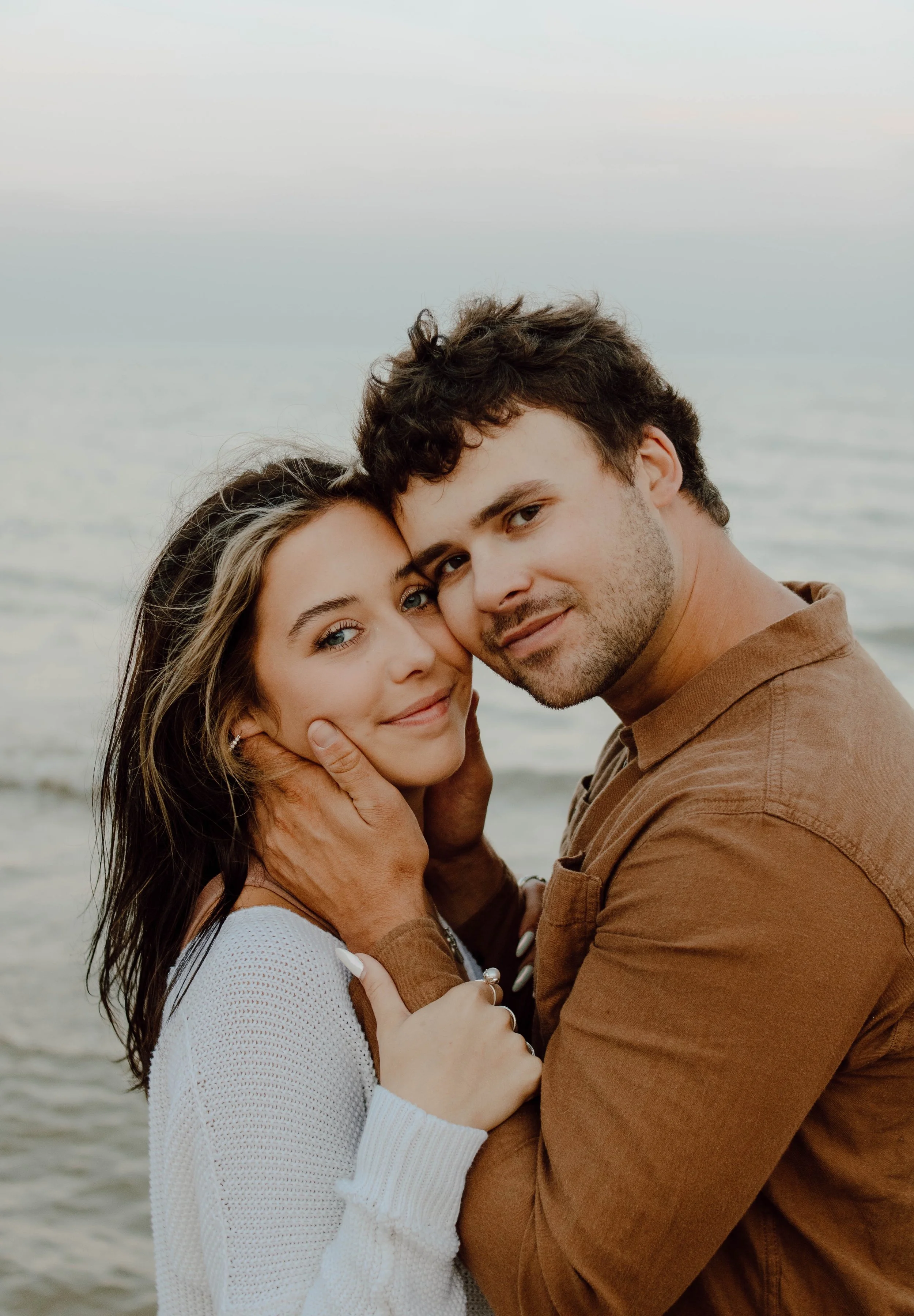 A young couple embraces by the sea, facing the camera with gentle smiles, during sunset.