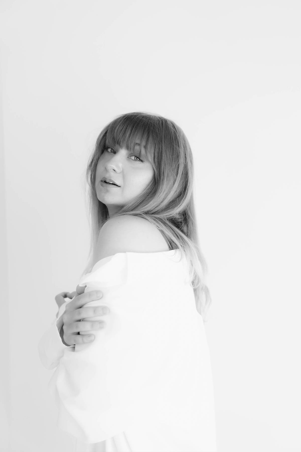 A black and white portrait of a woman with long hair and bangs, looking over her shoulder with her arms crossed, against a plain white background.