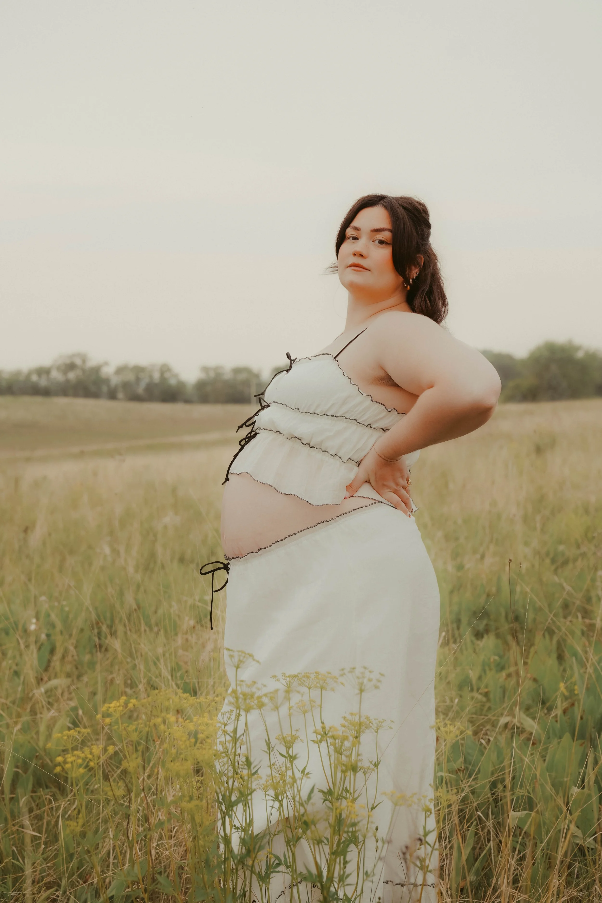 A pregnant woman standing in a grassy field, wearing a tied, ruffled, black and white dress, with her hand on her hip and looking confidently at the camera.