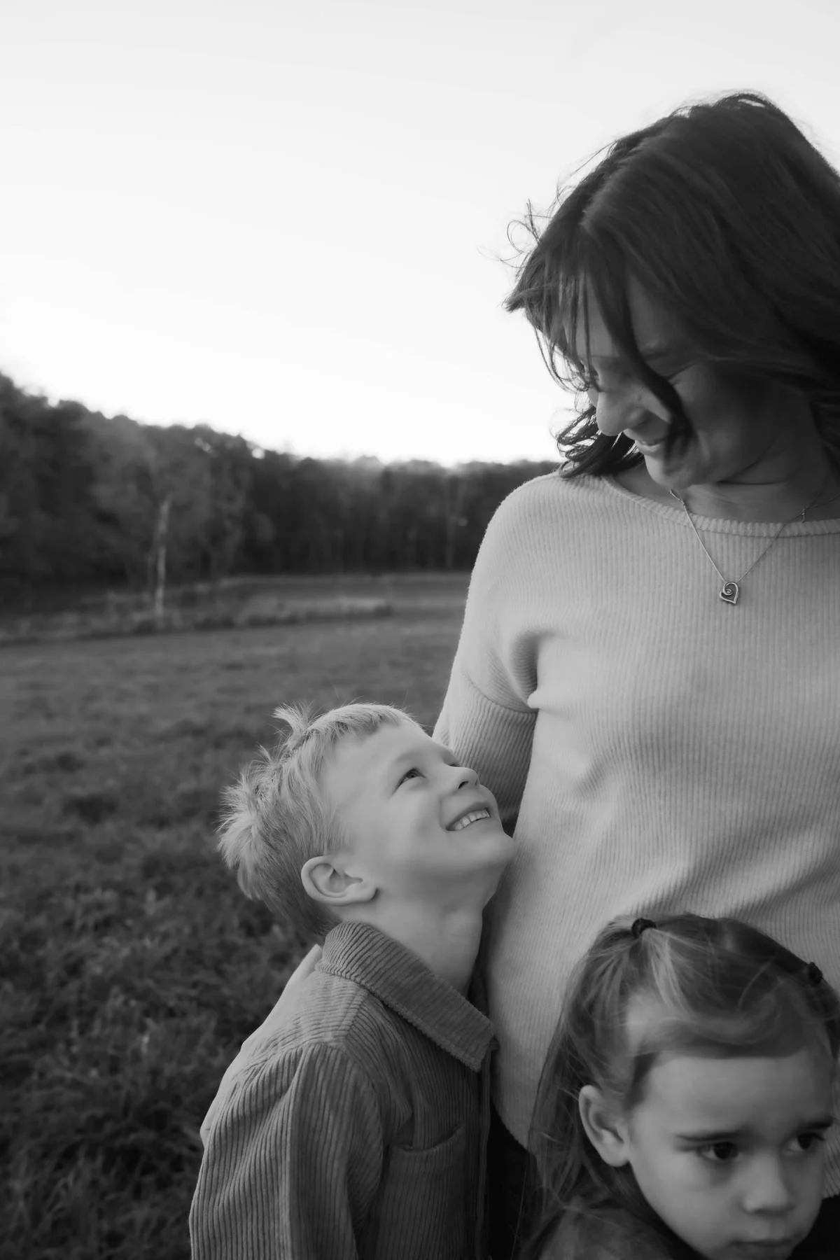 A woman smiling at a young boy who looks up at her lovingly, with a girl partially visible in the foreground, outdoors in a grassy field with trees in the background.