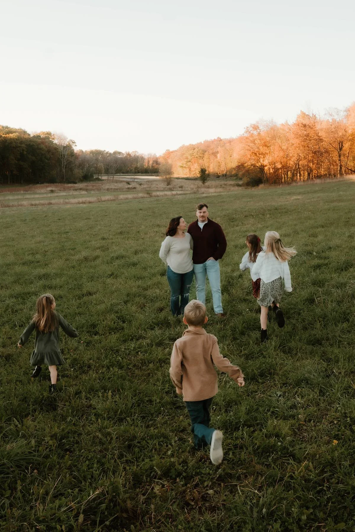 A family of six, including two adults and four children, walking in a grassy field during sunset with trees in the background.
