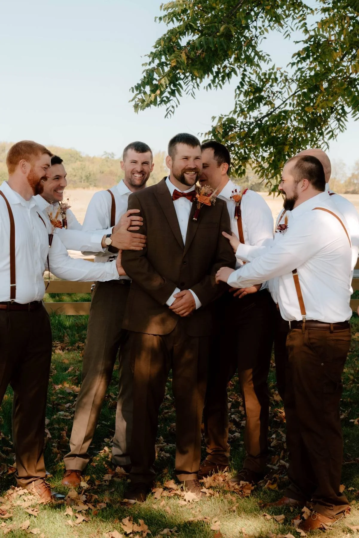 A groom surrounded by six groomsmen outdoors on a sunny day, with trees and a fence in the background.