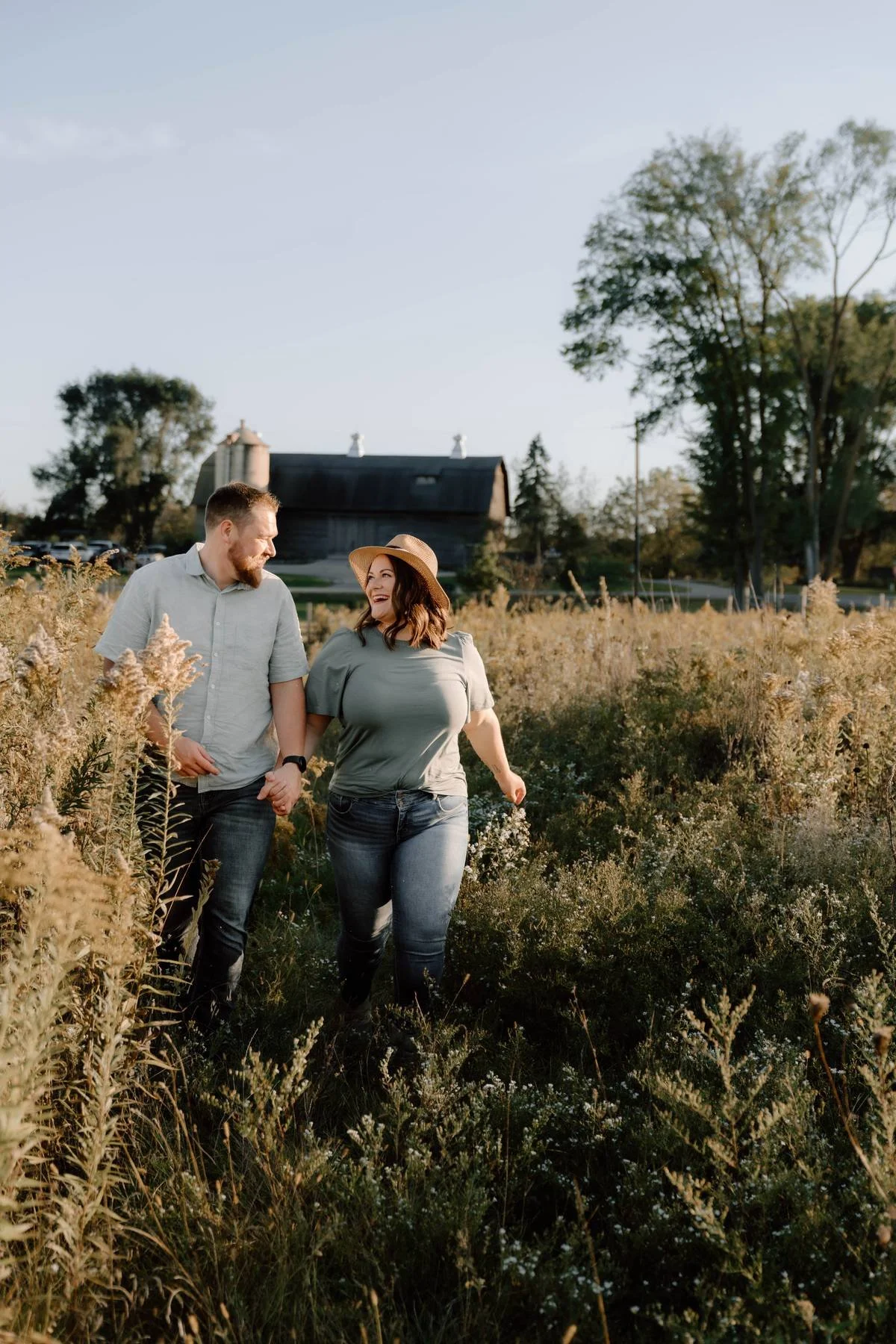 A couple walking through a field of tall grasses and wildflowers, holding hands and smiling at each other, with a barn and trees in the background.