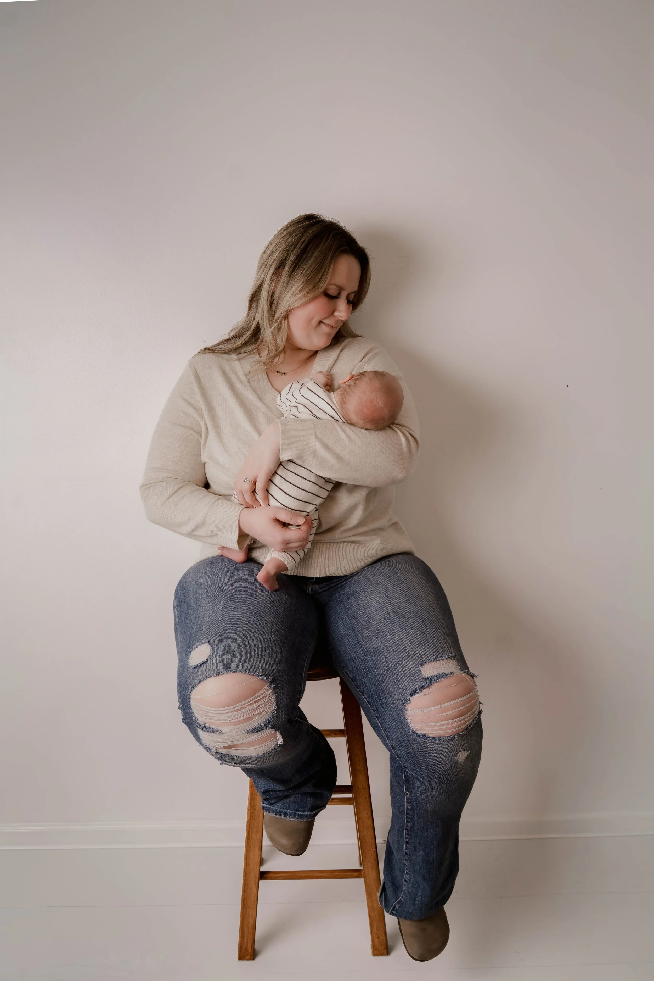 A woman sitting on a wooden stool holding a newborn baby in her arms, both facing each other and looking at the baby, in a simple room with white walls.