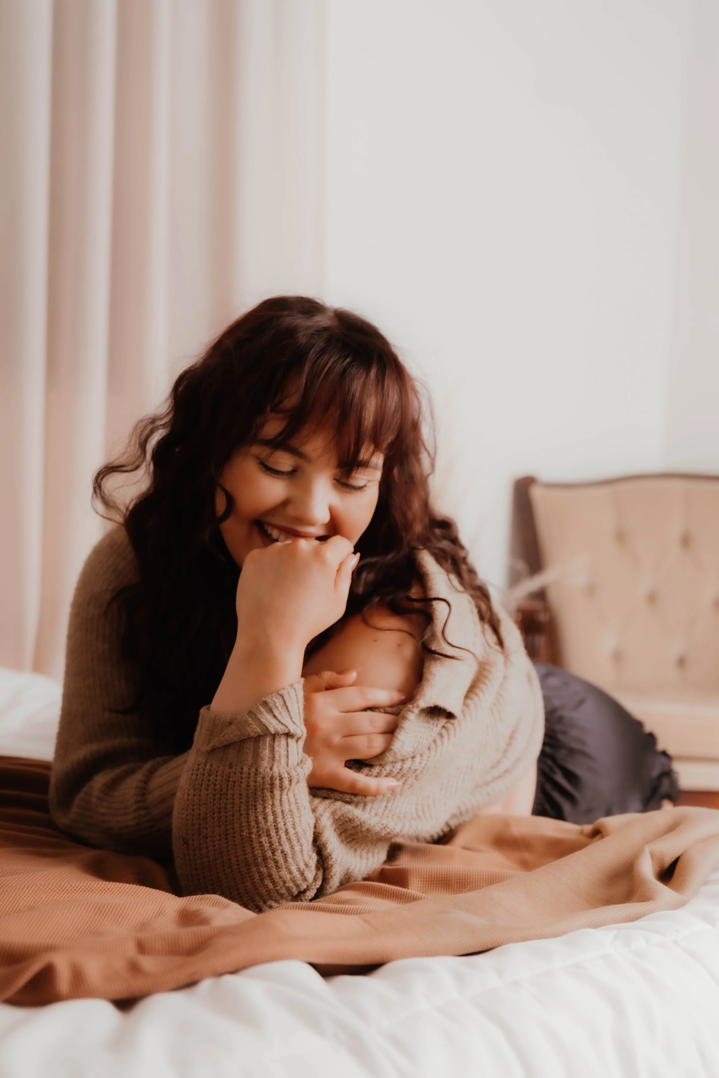 A woman with curly brown hair smiling while lying on a bed, cradling a baby wrapped in a beige blanket.