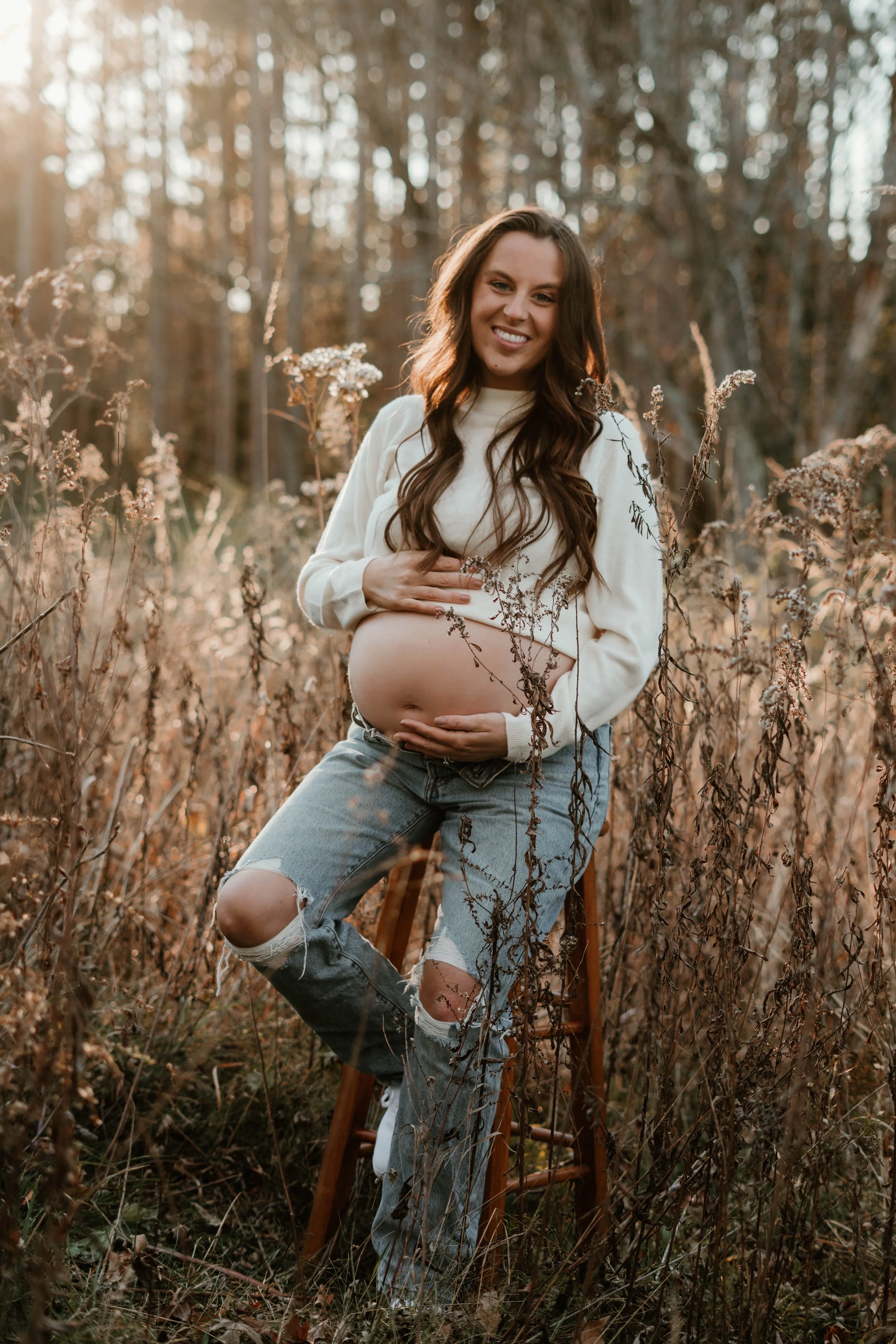 Pregnant woman sitting on a wooden stool in a field of tall, dried grass and plants, smiling at the camera during sunset, with trees in the background.