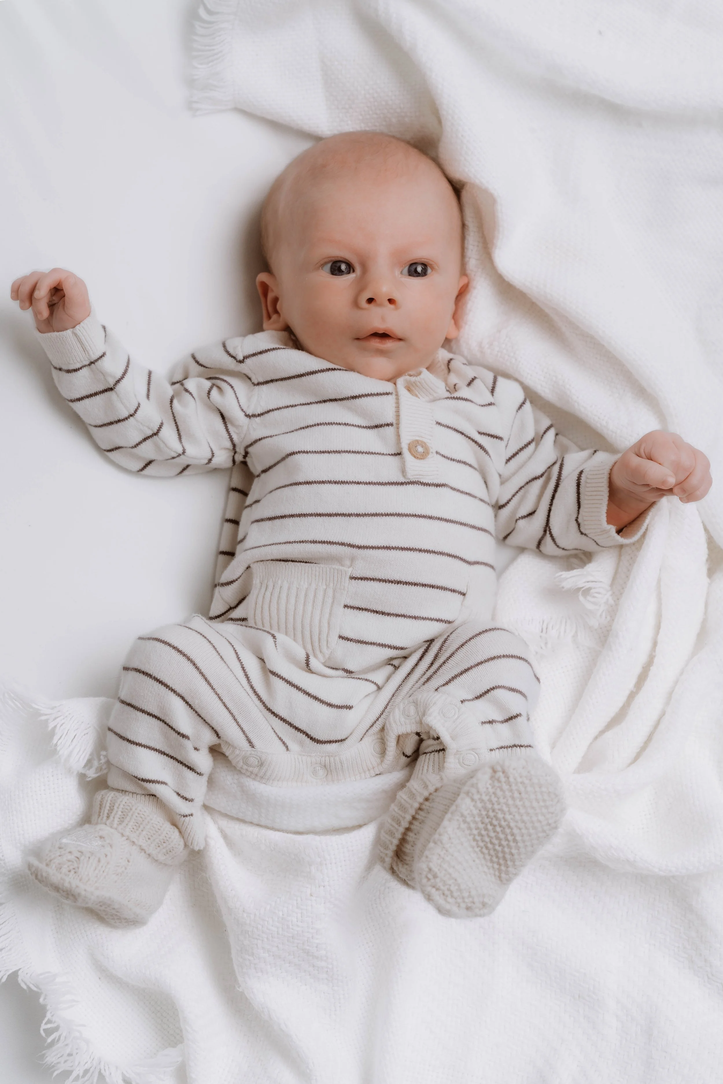 A baby in a striped onesie lying on a white blanket.