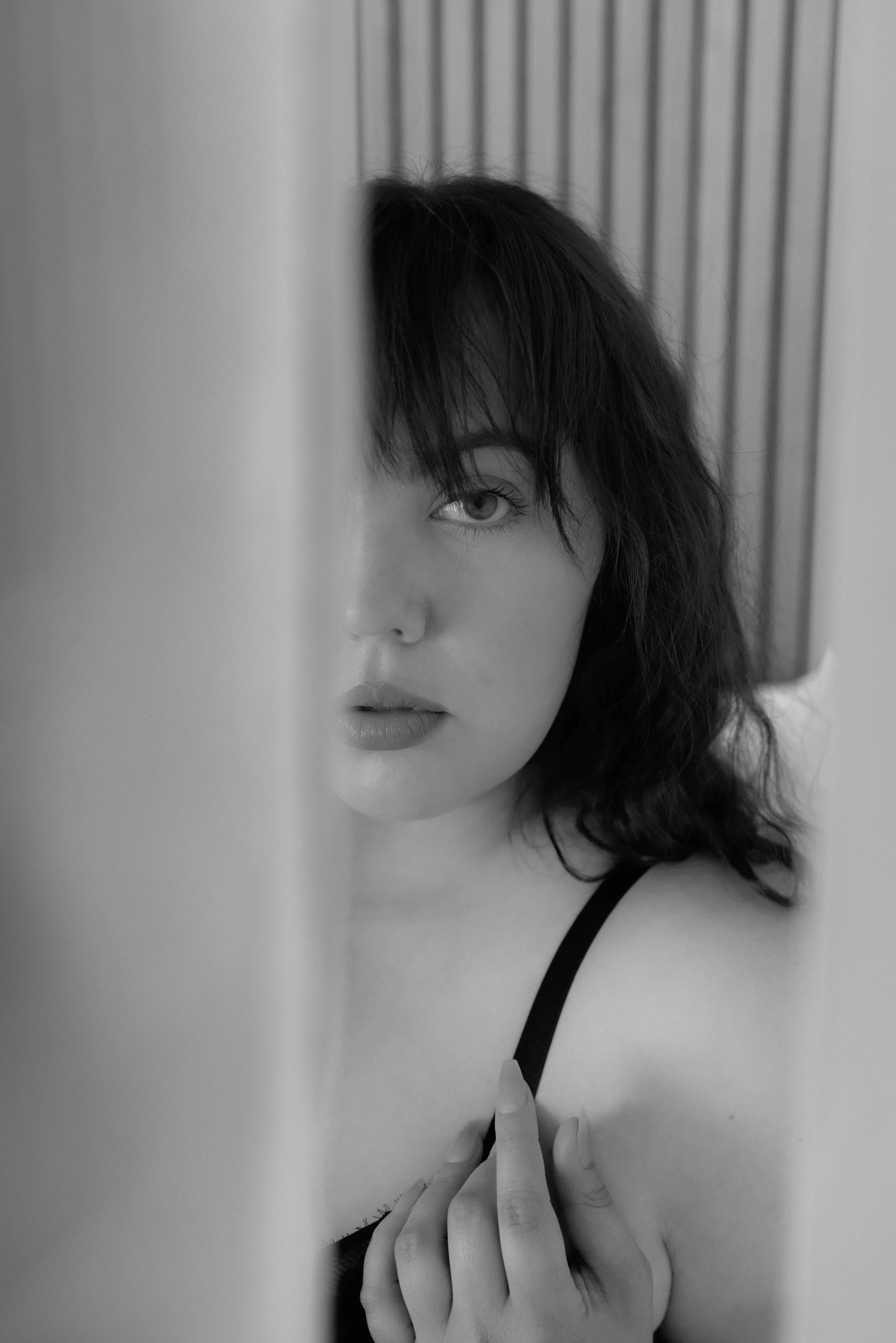 A black and white photo of a woman with dark hair, visible eye, and partially covered face, standing near a wall or panel.