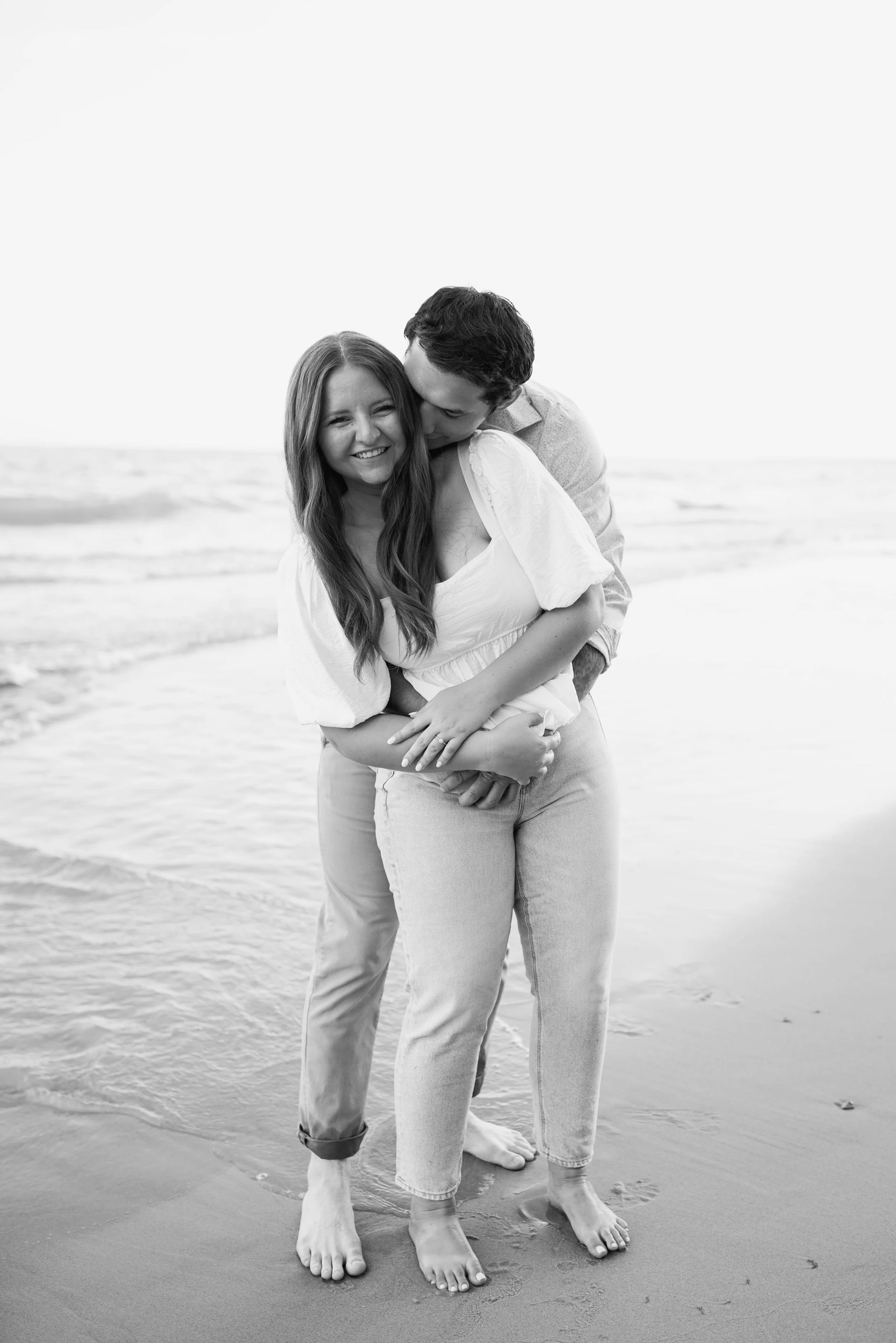 A black and white photo of a smiling woman and a man on a beach, with the man hugging the woman from behind, both barefoot and standing on wet sand near the ocean.