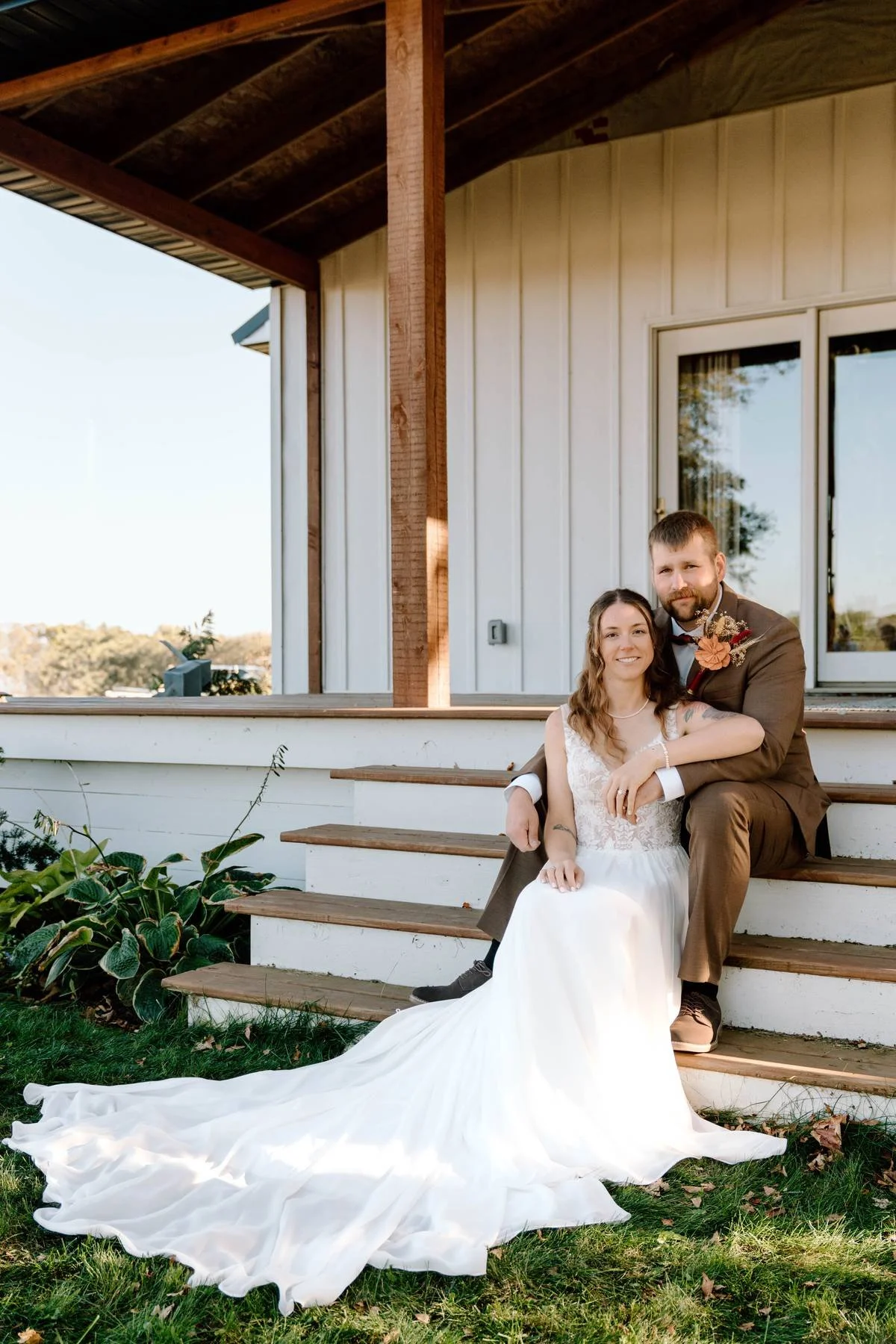 A bride and groom sitting on outdoor wooden steps. The bride wears a white wedding dress, and the groom wears a brown suit with boutonniere. They are smiling, with the bride resting her arm on the groom's shoulder. Green foliage and a grassy area are
