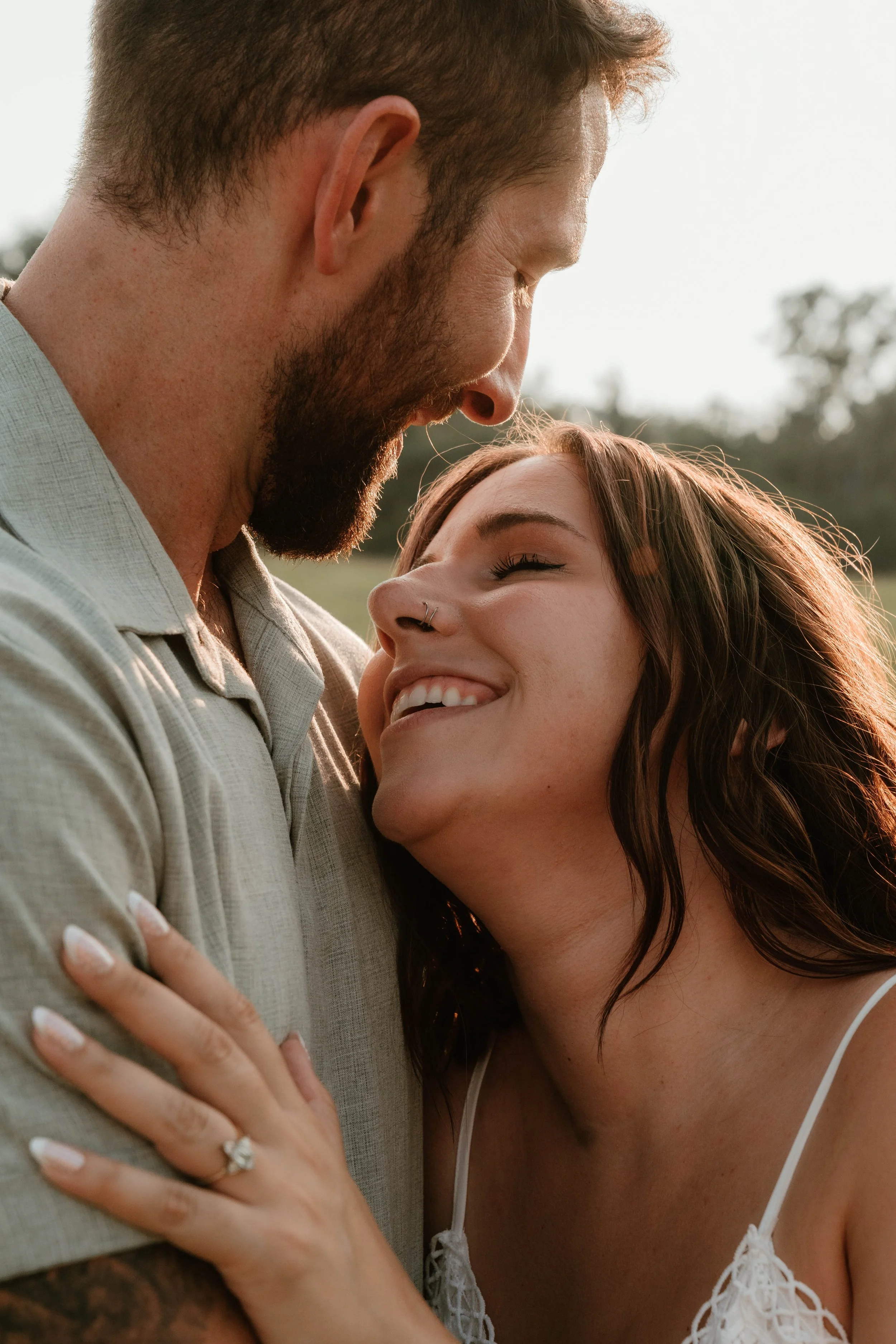 A couple sharing a tender moment outdoors, close-up, with the woman smiling and the man gently leaning his forehead to hers.