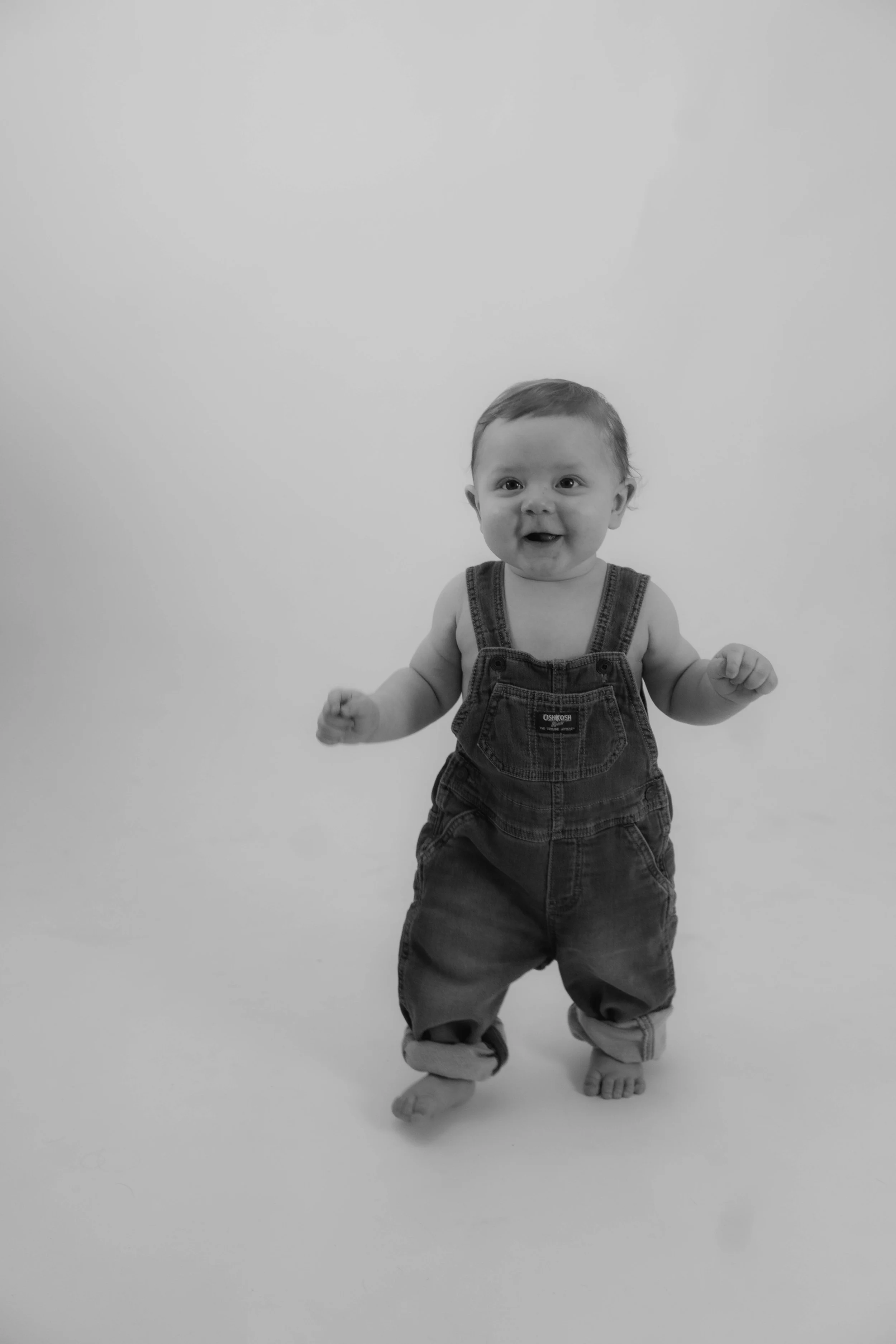 Black and white photo of a baby standing and smiling, wearing overalls with rolled-up pant legs.