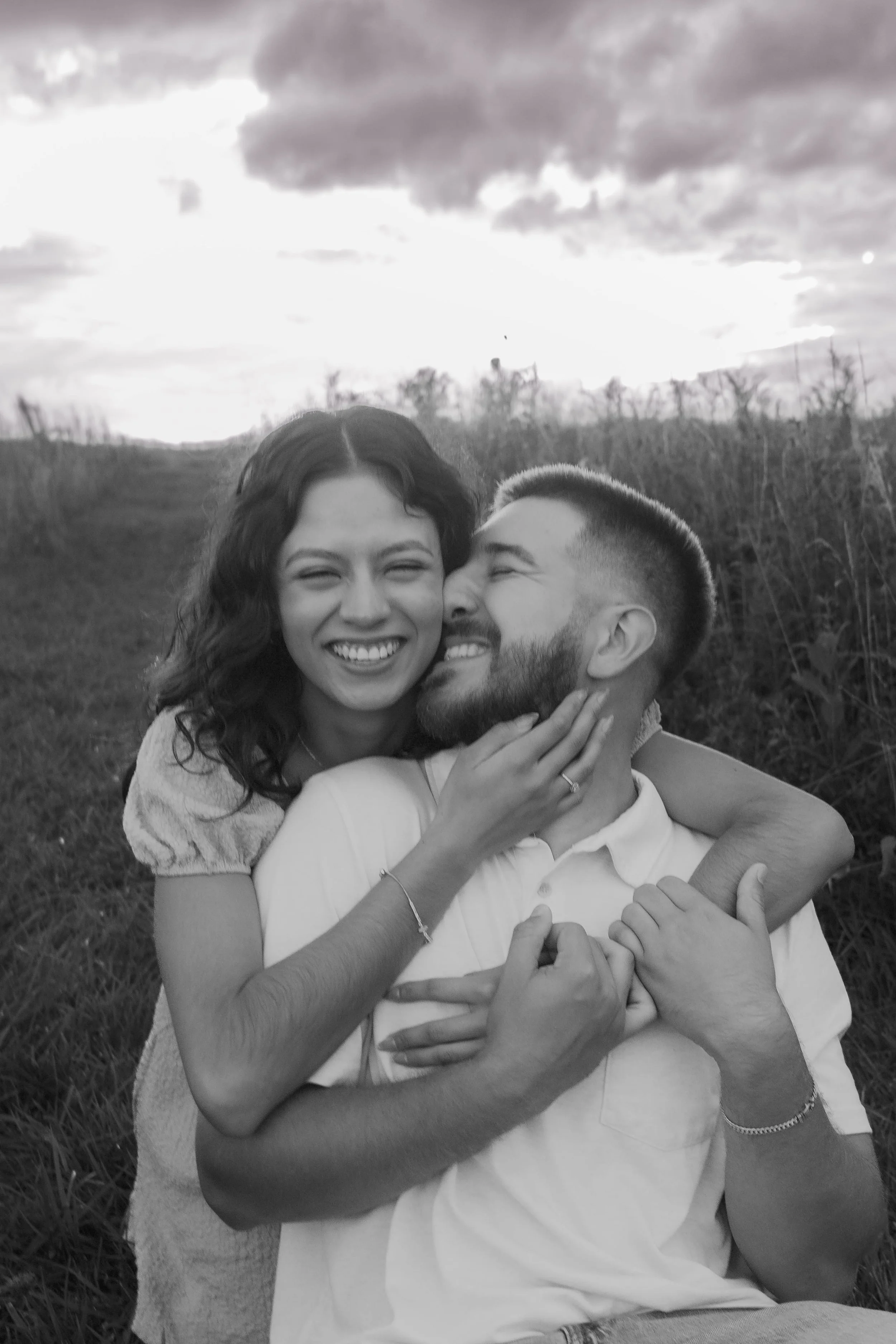 A happy couple appears to be playing together outside at sunset, with cloudy sky and grassy field in the background. The woman is smiling and holding the man’s face, while the man is smiling and embracing her.